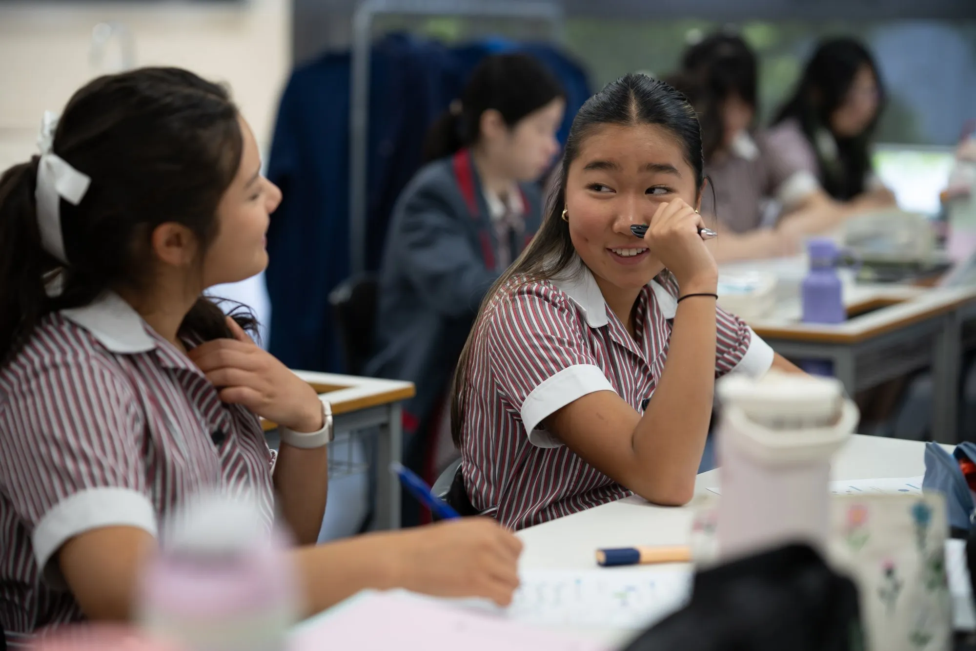 Two female students in striped uniforms sitting at desks, smiling and talking in a classroom.
