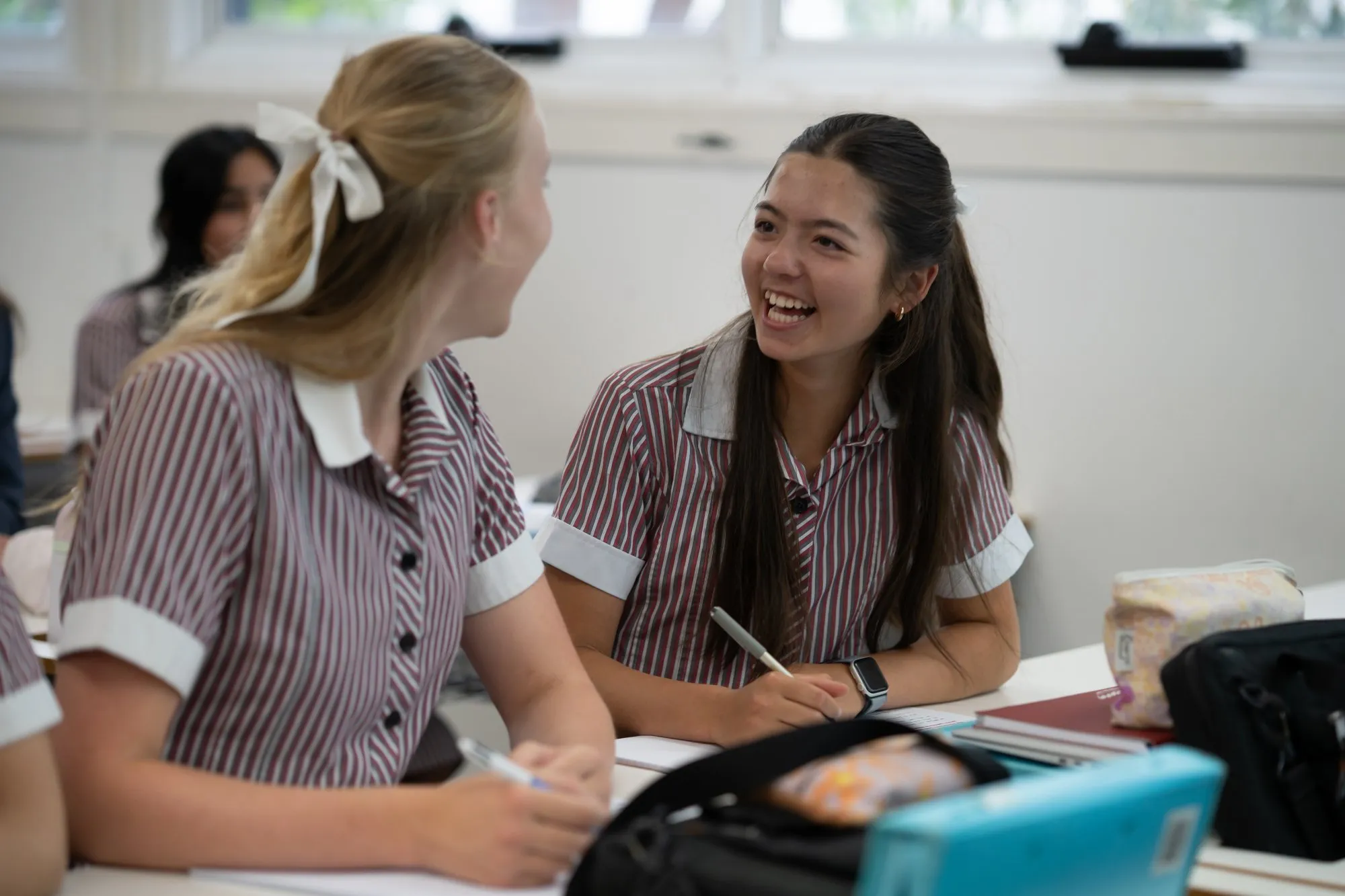 Two female students in striped uniforms smiling and talking in a classroom while writing in notebooks.