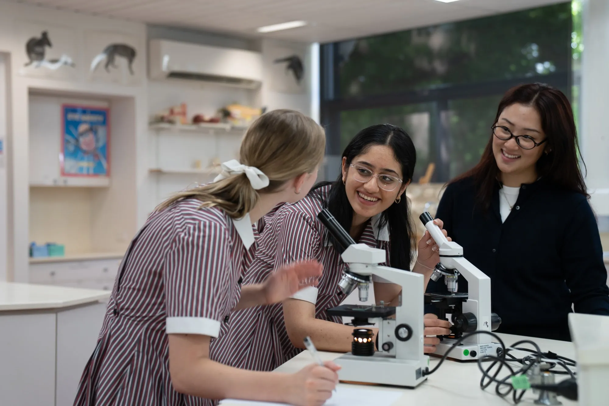 Three female students smiling and using microscopes in a science classroom.