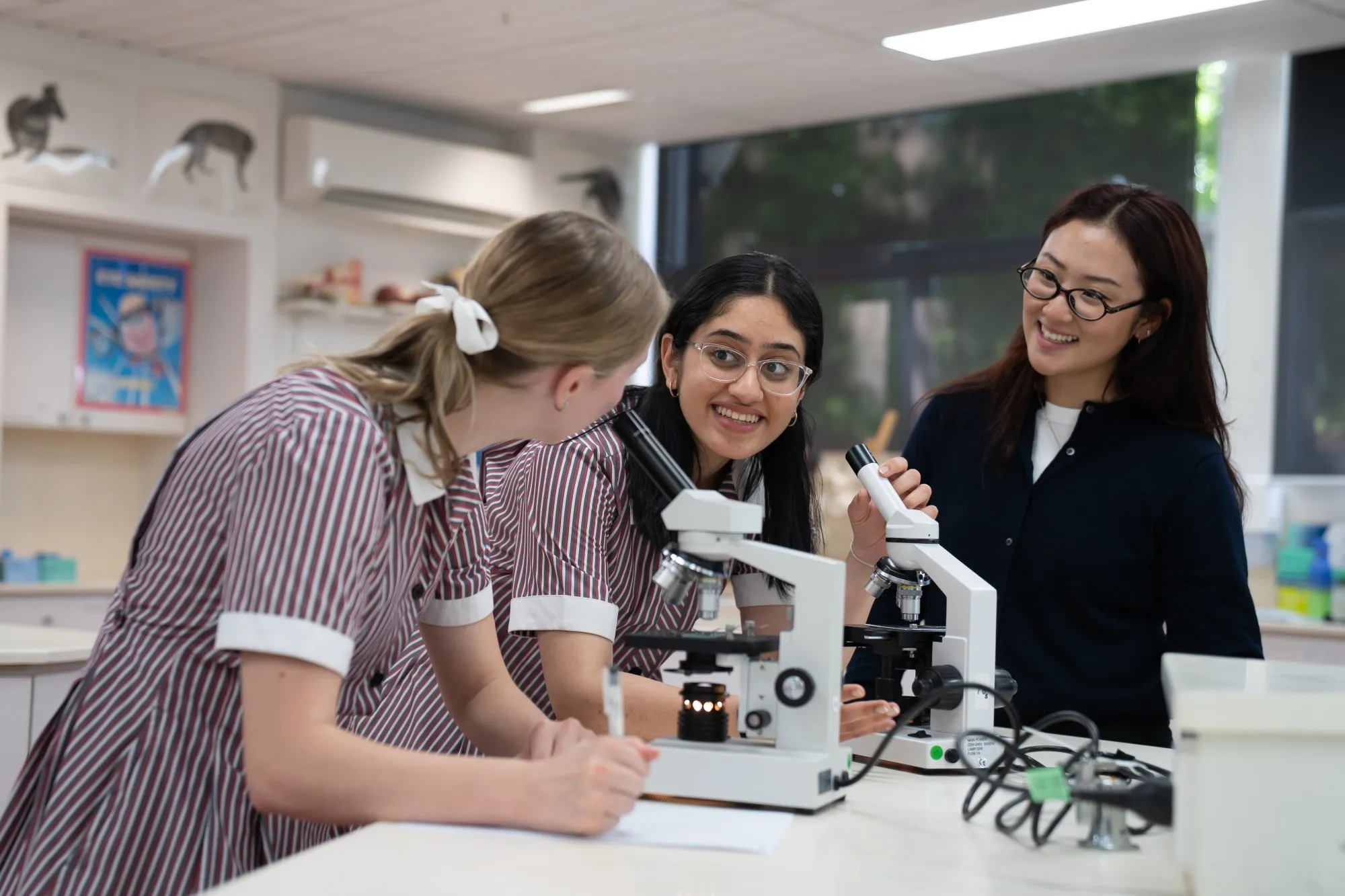 Three female students in a science lab, two using microscopes and one taking notes, smiling and interacting.