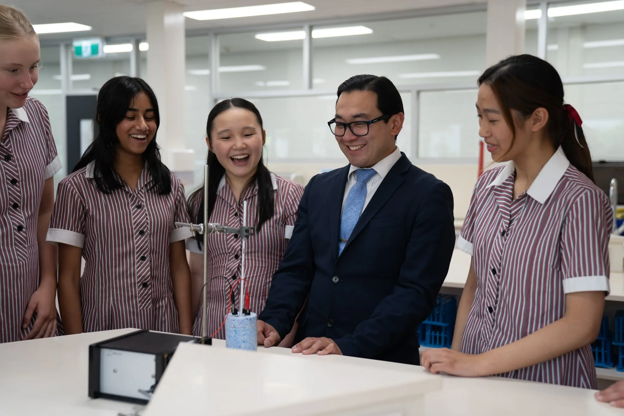 A group of high school students in striped uniforms and a teacher in a suit smile while observing a science experiment in a laboratory.