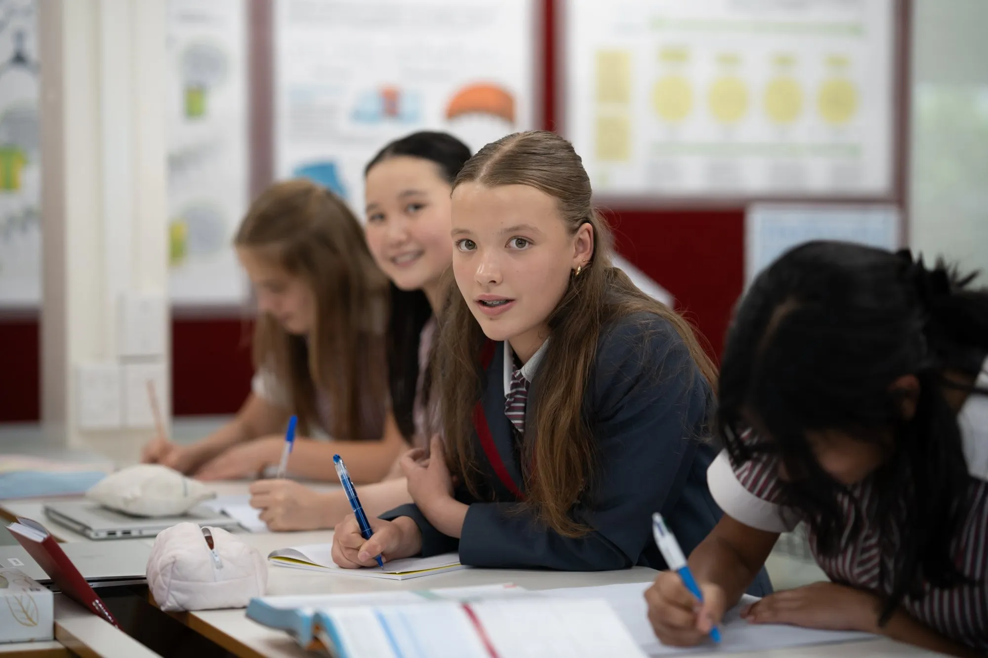 Four girls in school uniforms sitting at a desk writing in notebooks, with one girl looking directly at the camera.