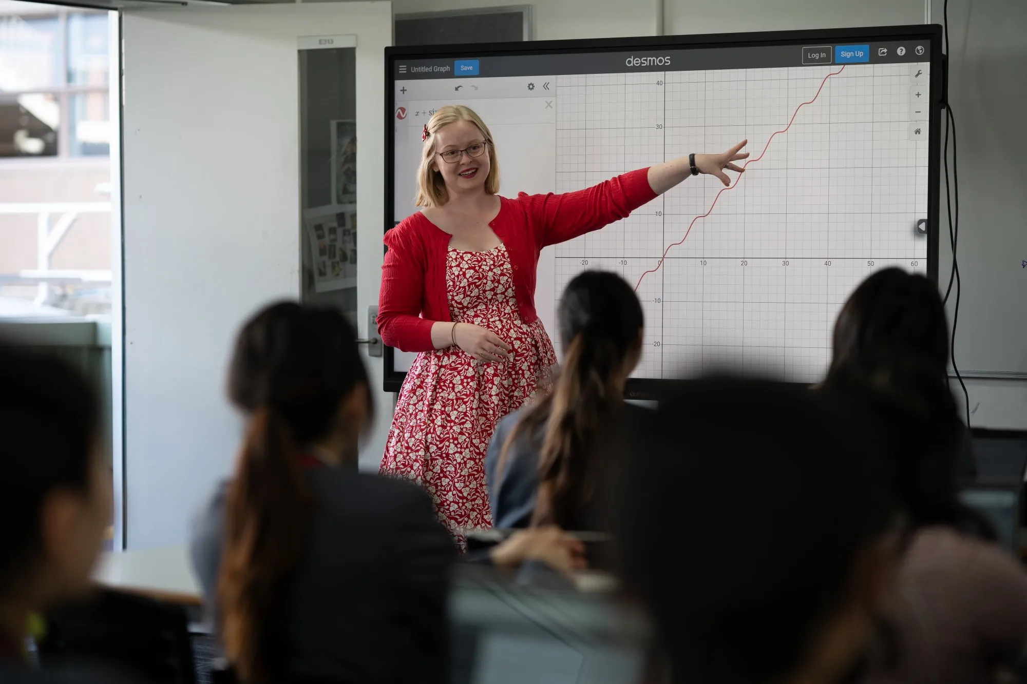 Female teacher in a red dress pointing at a rising graph on a large screen while students watch.