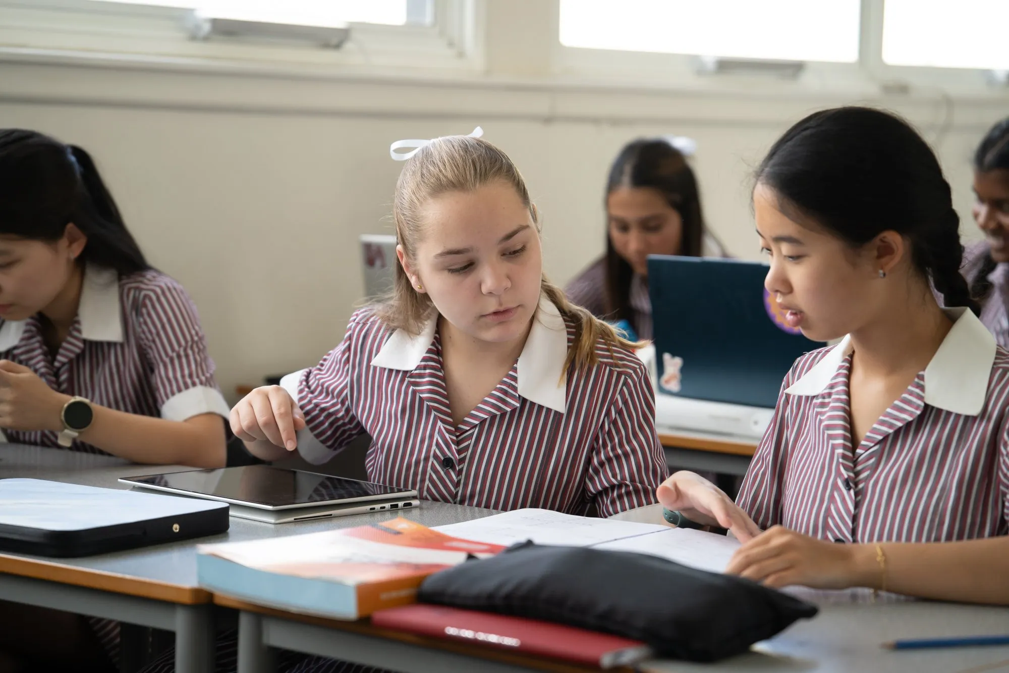 Two female students in striped uniforms studying together at a desk with a tablet and open notebook.