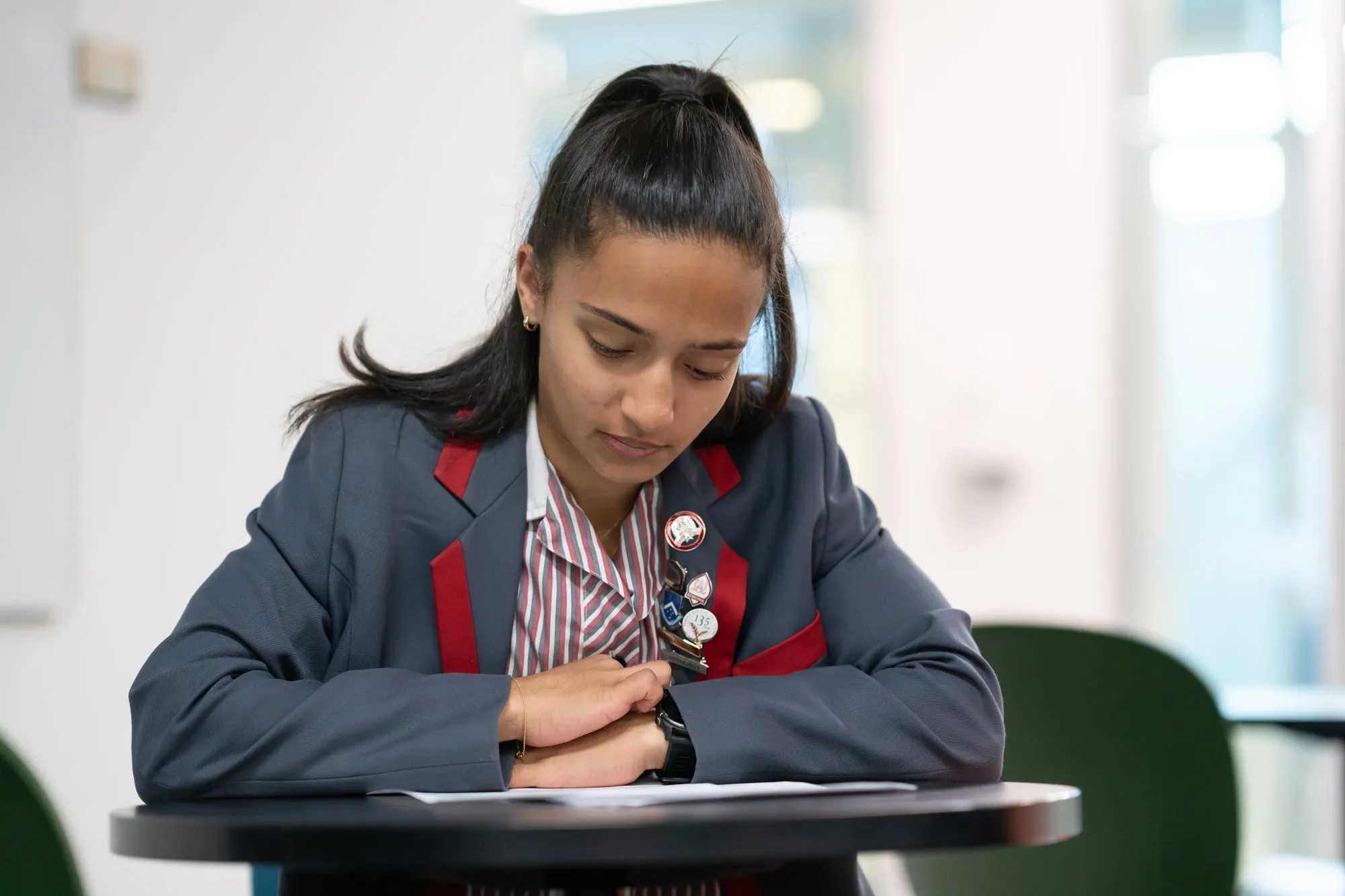 Young female student in school uniform reading a paper at a round table indoors.