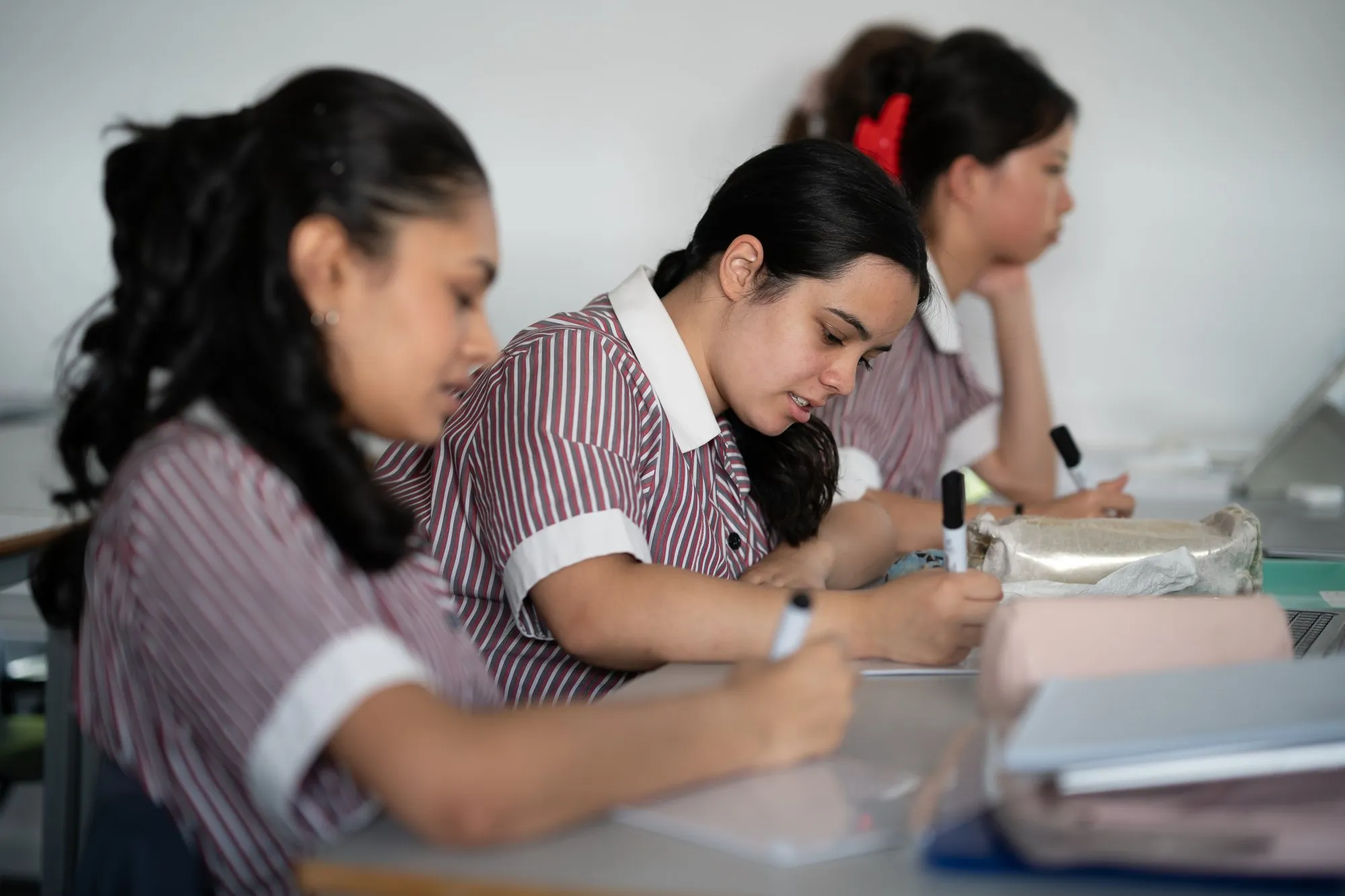 Three female students in striped uniforms sitting at desks, writing with markers in a classroom.