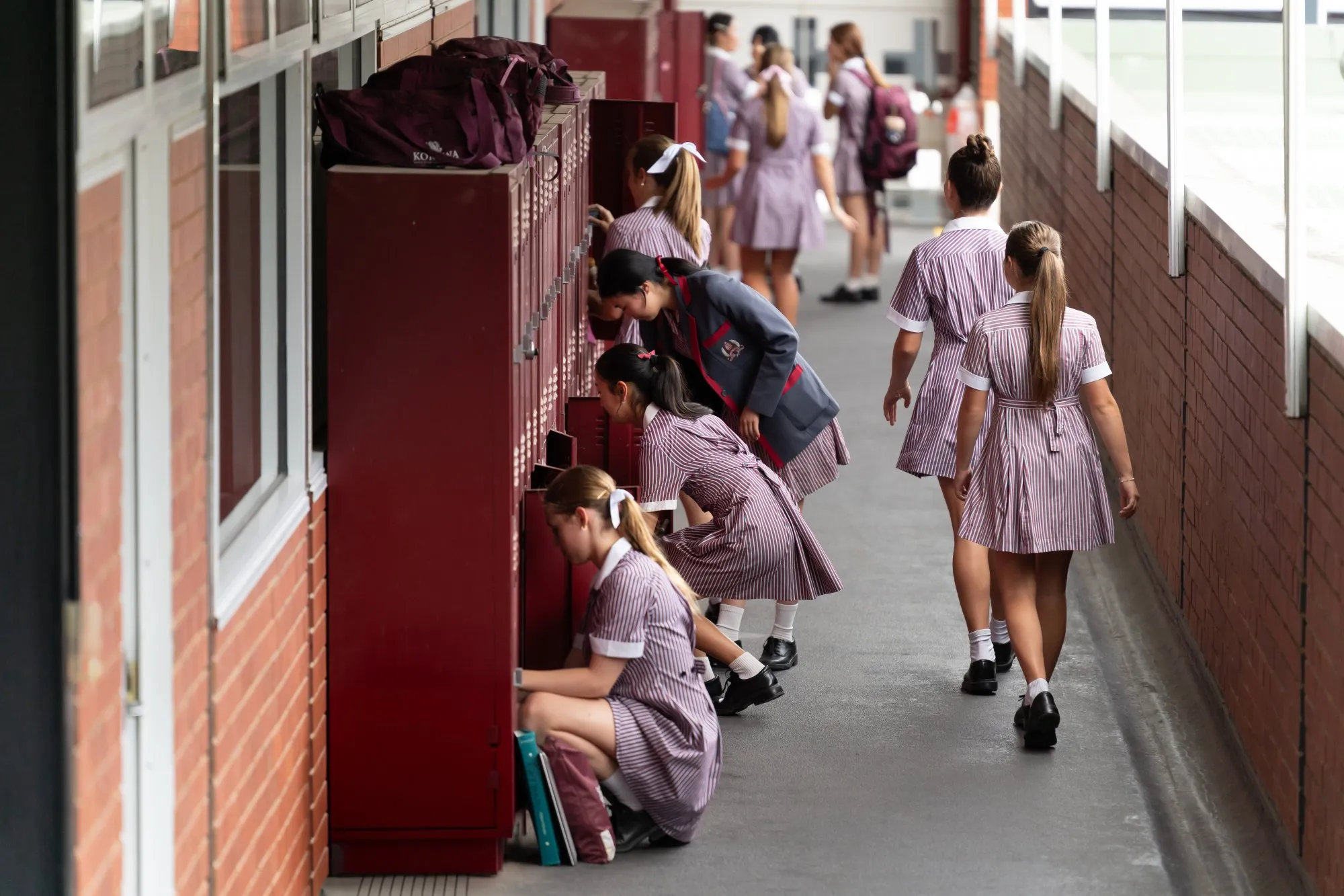 Schoolgirls in striped uniforms accessing lockers in a hallway, some are walking while others are opening or using their lockers.
