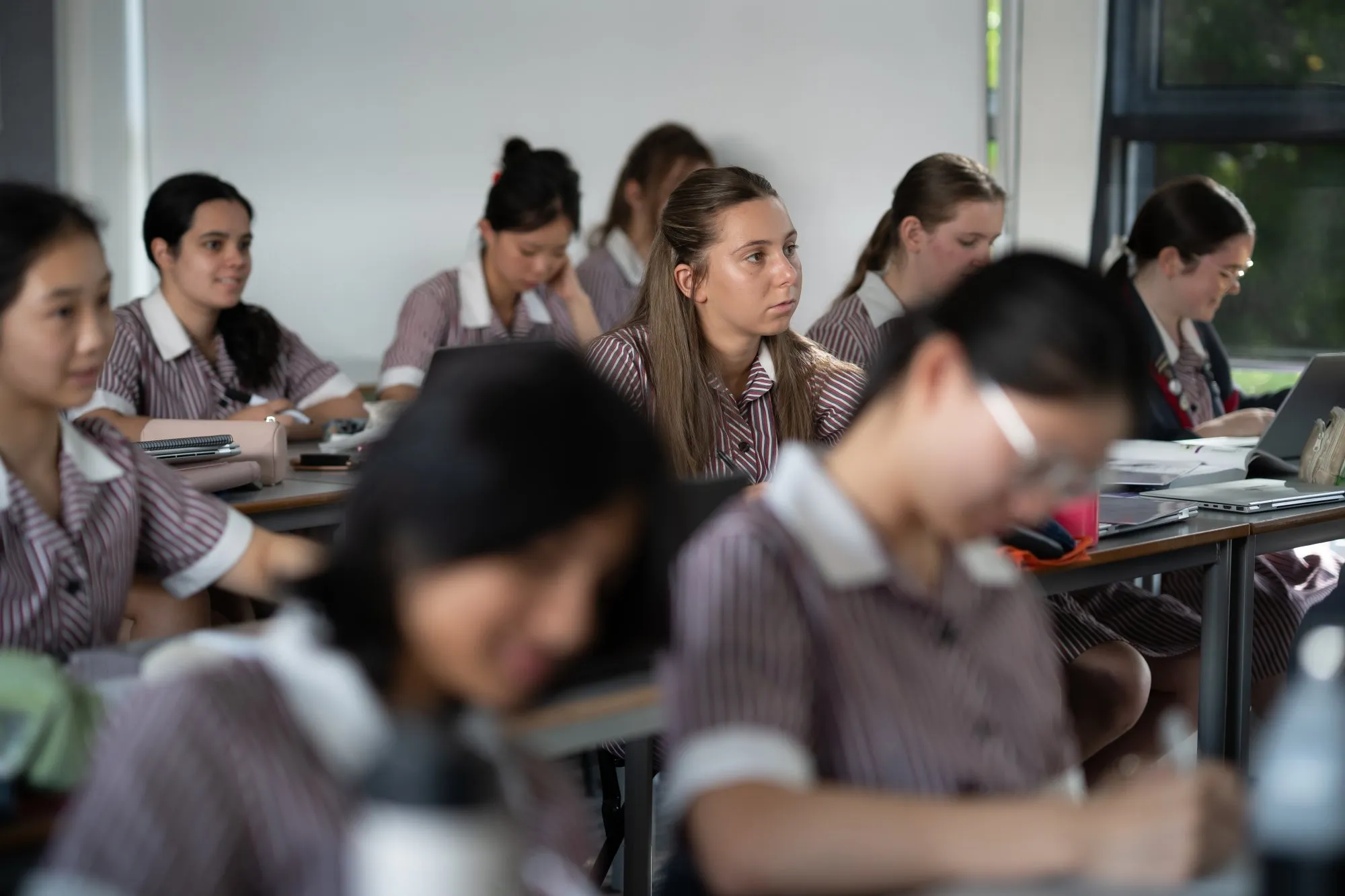 Female students in striped school uniforms seated in a classroom, some focused on their work and one looking attentively to the side.