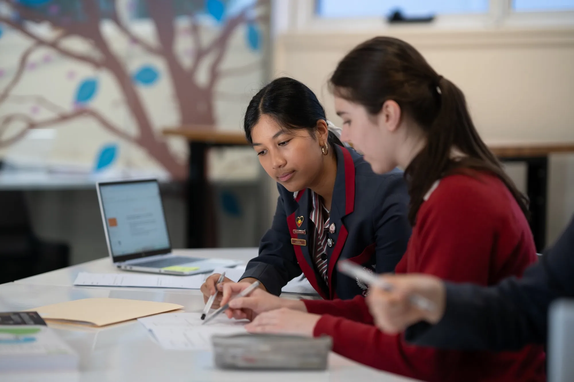 Two female students working together on papers at a table with a laptop in the background.