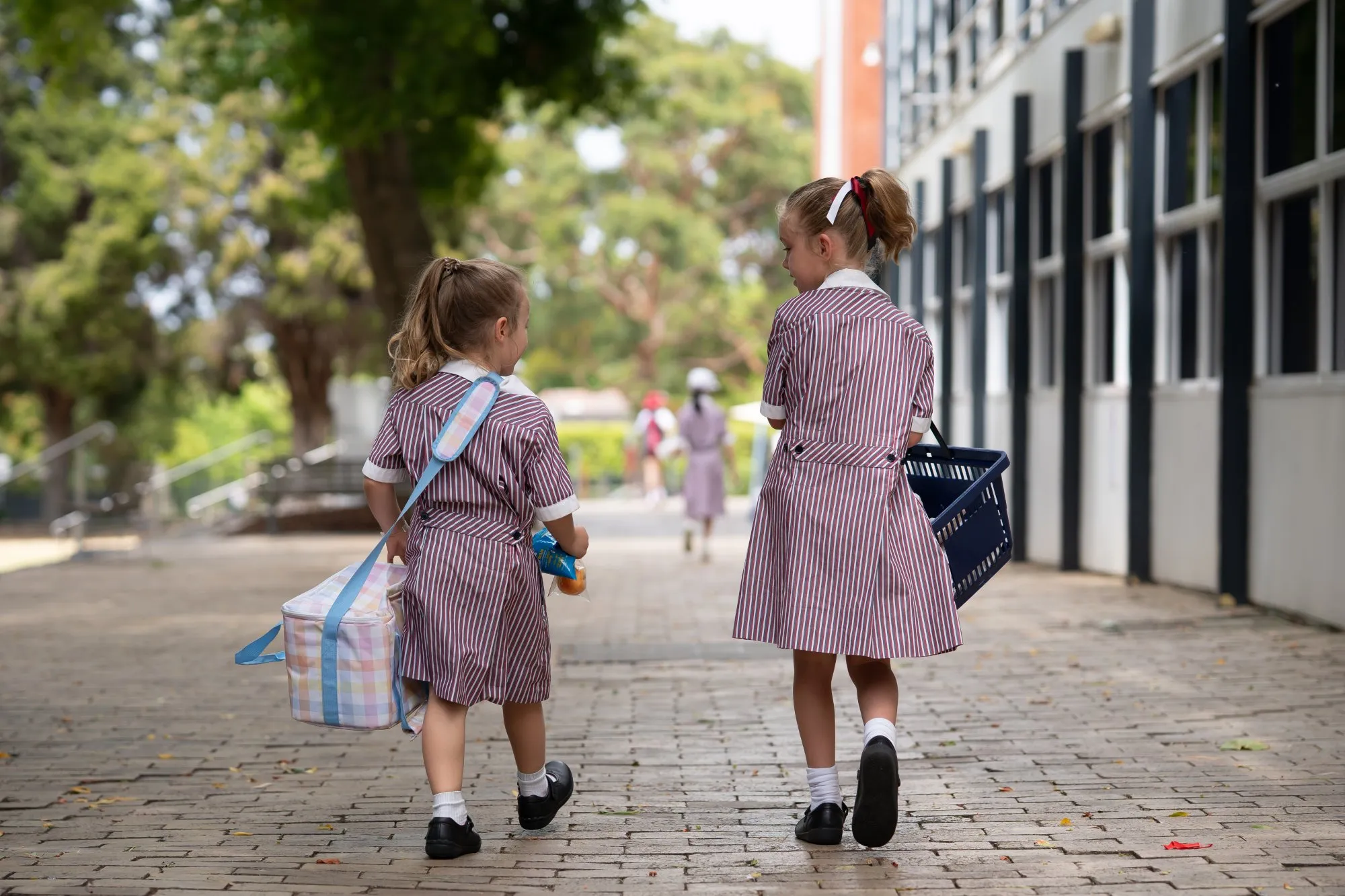 Two young girls in striped school uniforms walk on a brick pathway, carrying a lunch bag and a basket, with a school building on the right.