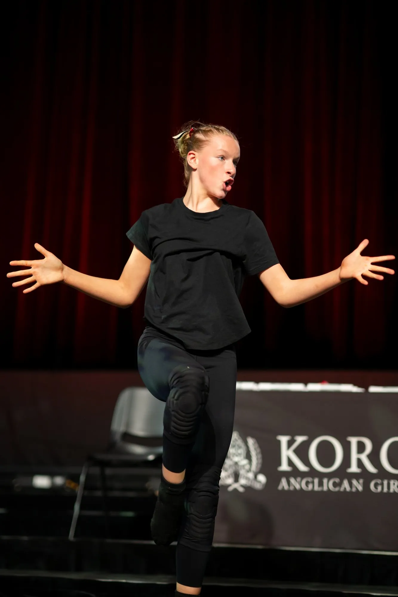 Young dancer in black outfit performing on stage with expressive hand gestures against a dark red curtain backdrop.