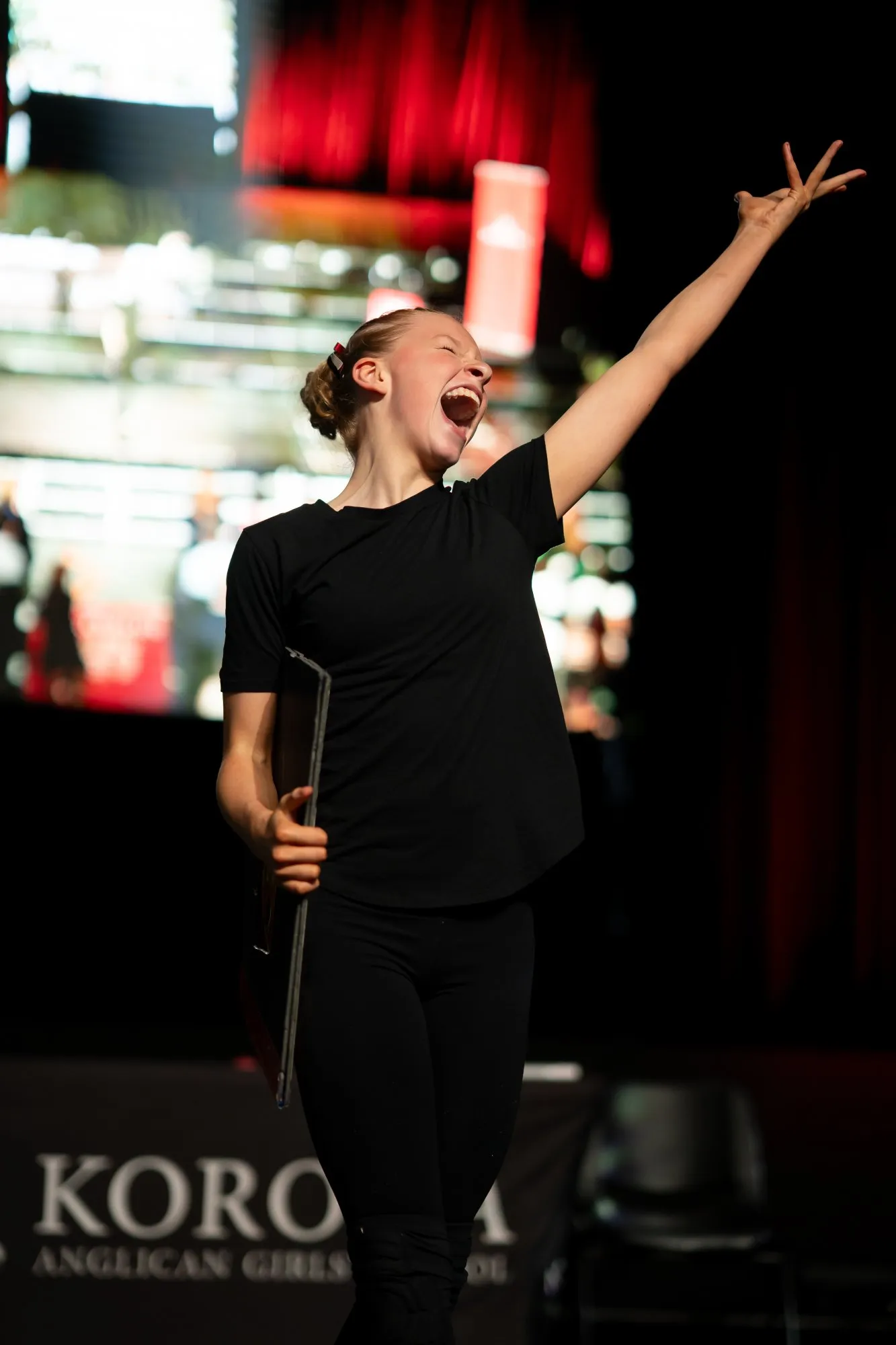 Young woman in black holding a folder, singing or shouting with one arm raised on stage.
