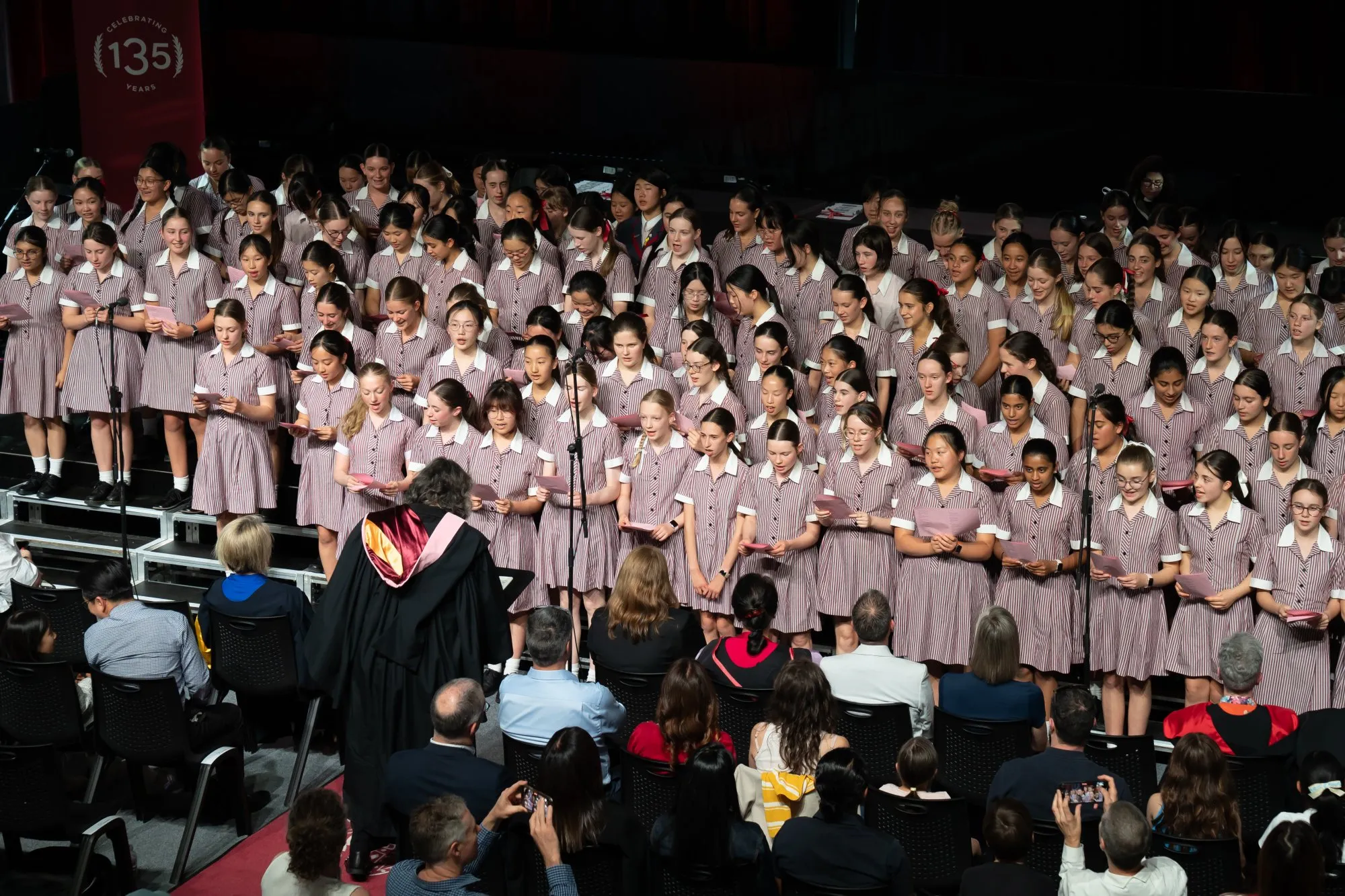 Large group of schoolgirls in striped uniforms singing on stage, directed by a conductor, with seated audience watching.