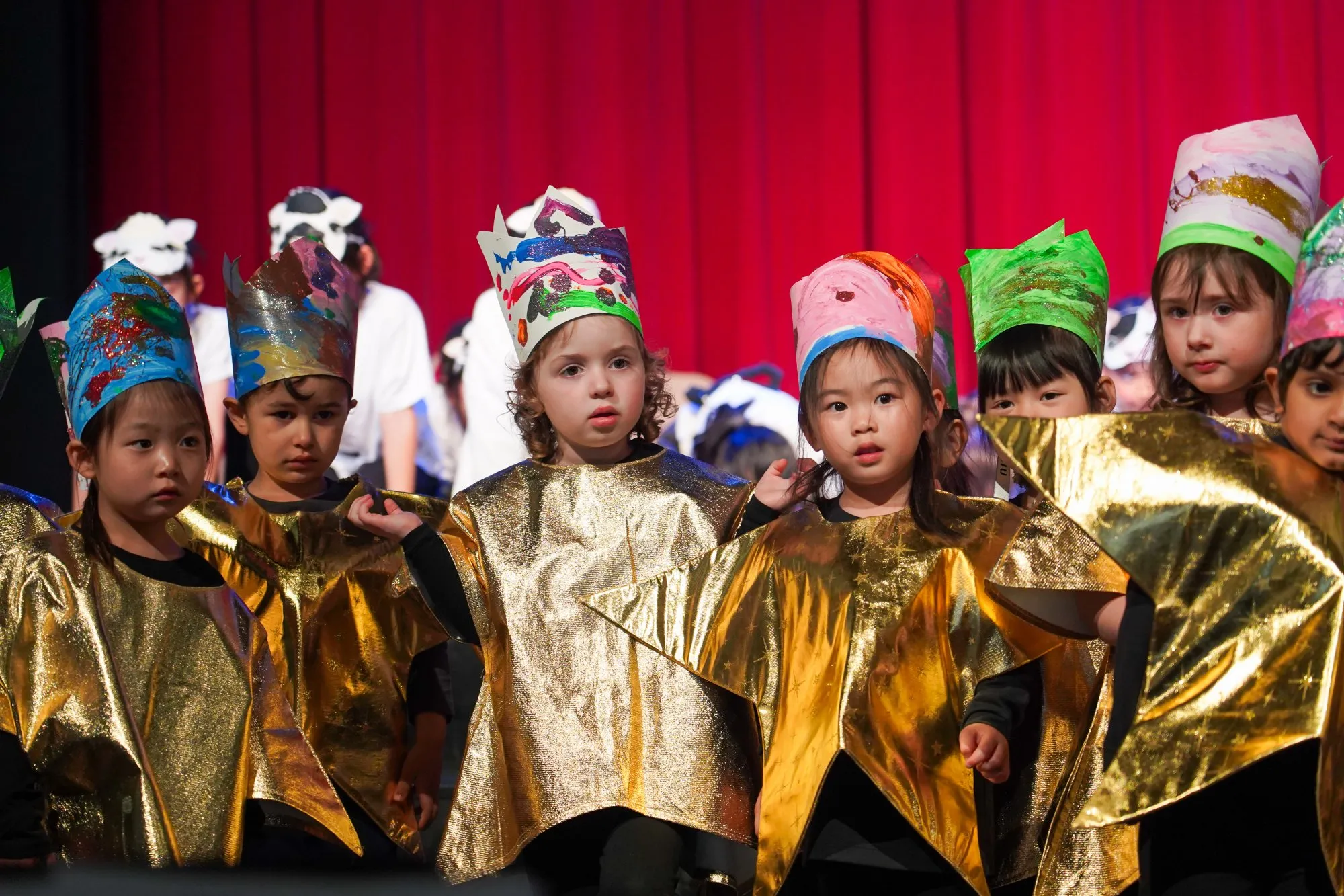 Group of young children wearing colorful handmade crowns and gold star costumes on stage against a red curtain.