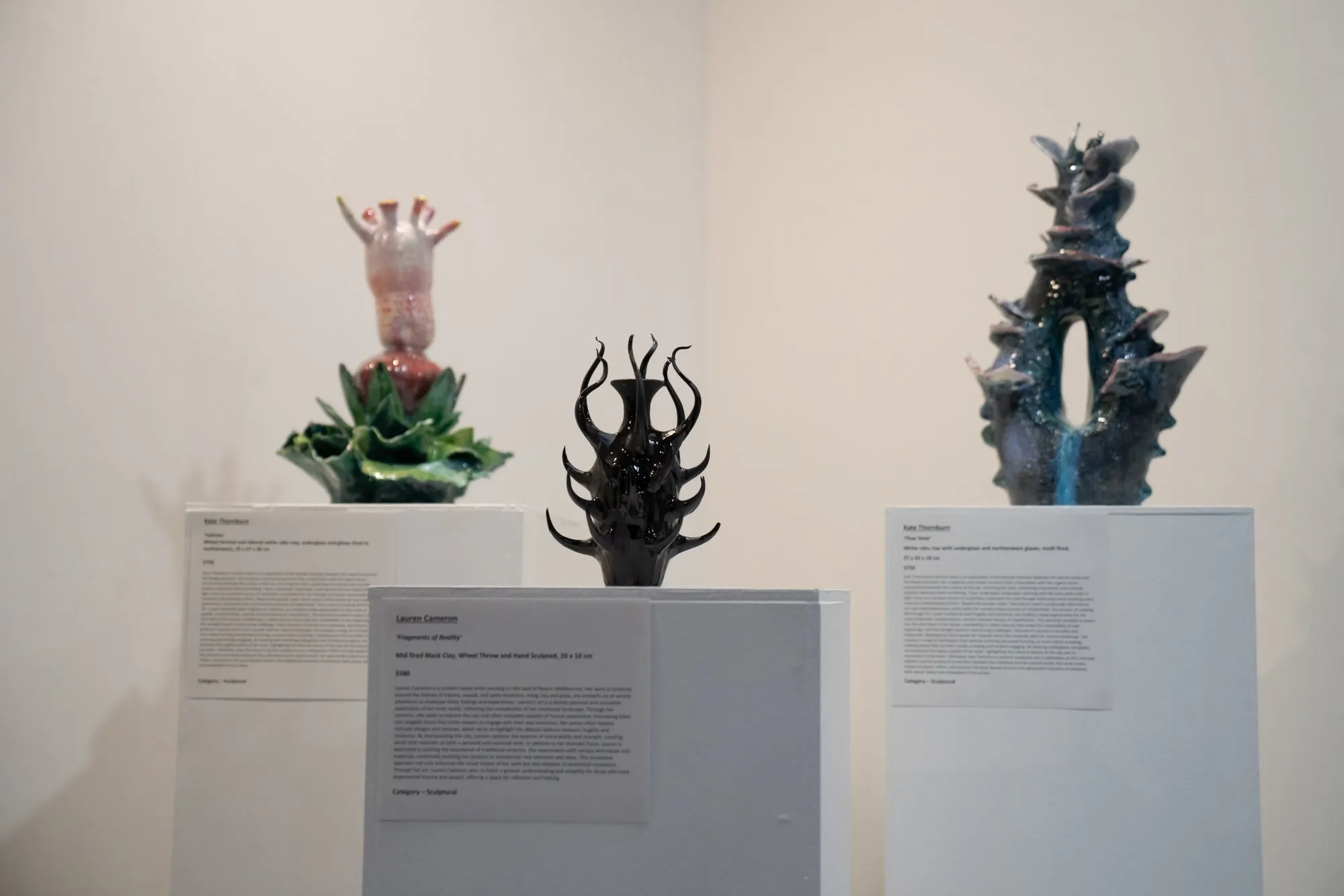 Three abstract ceramic sculptures displayed on white pedestals in a gallery setting.