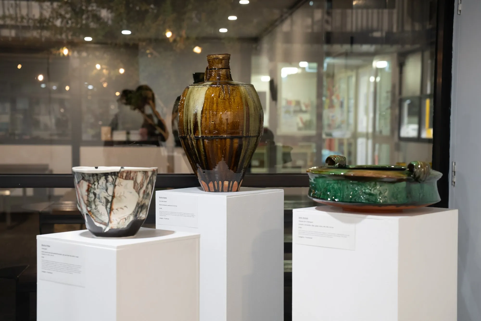 Three ceramic art pieces displayed on white pedestals in a gallery setting.