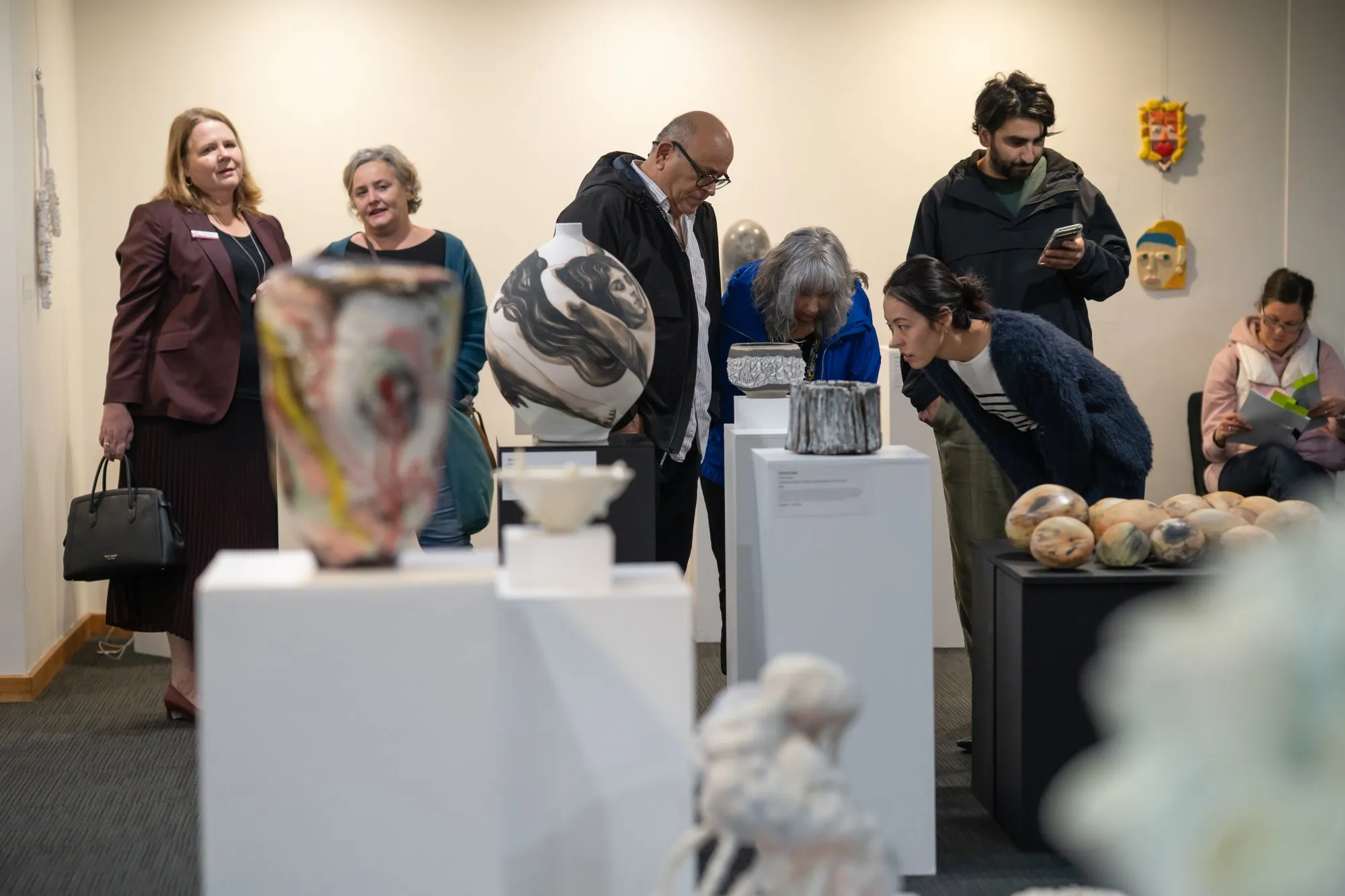 Group of people observing ceramic art pieces displayed on white pedestals in an art gallery.