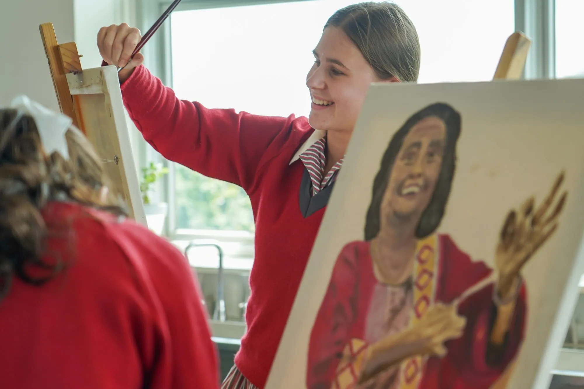 Smiling girl in a red sweater painting on a canvas in a bright room.