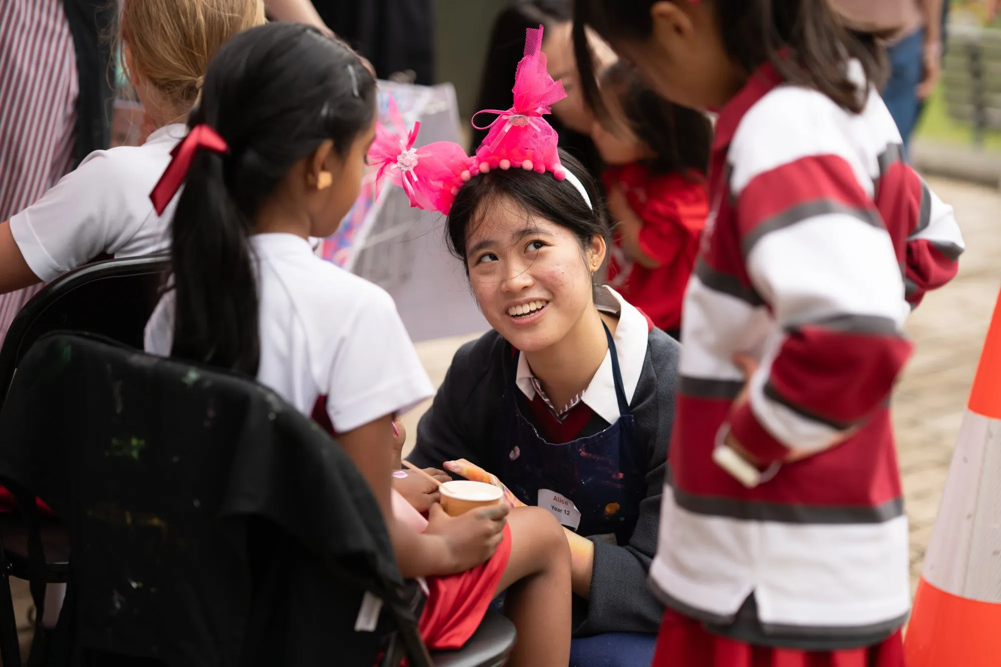 A young woman wearing a pink bow headband smiles while interacting with children at a seated activity.