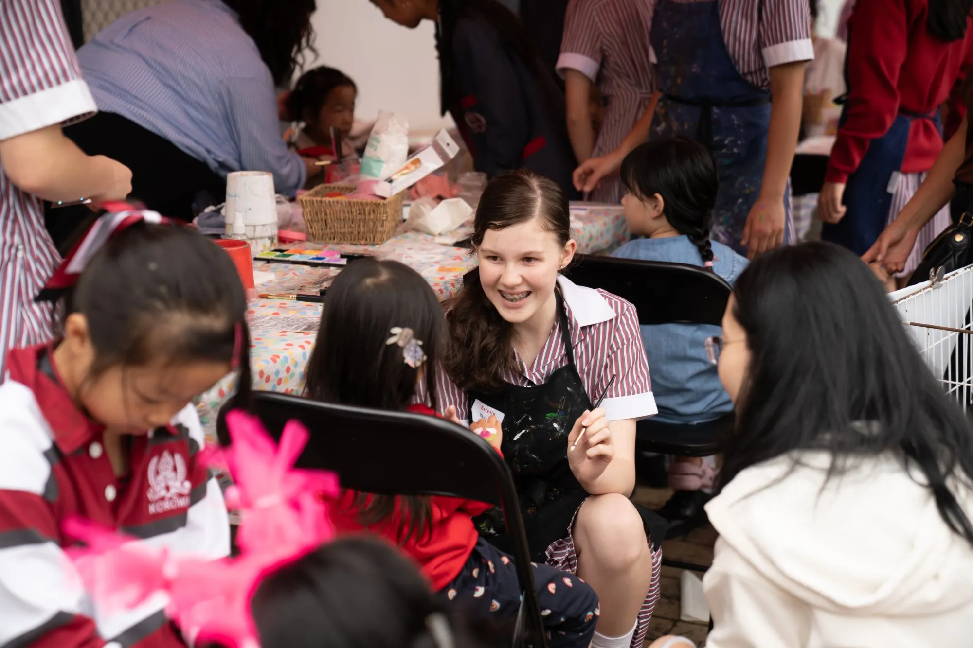 A smiling young girl in a striped uniform and black apron interacts with children at a craft activity table.