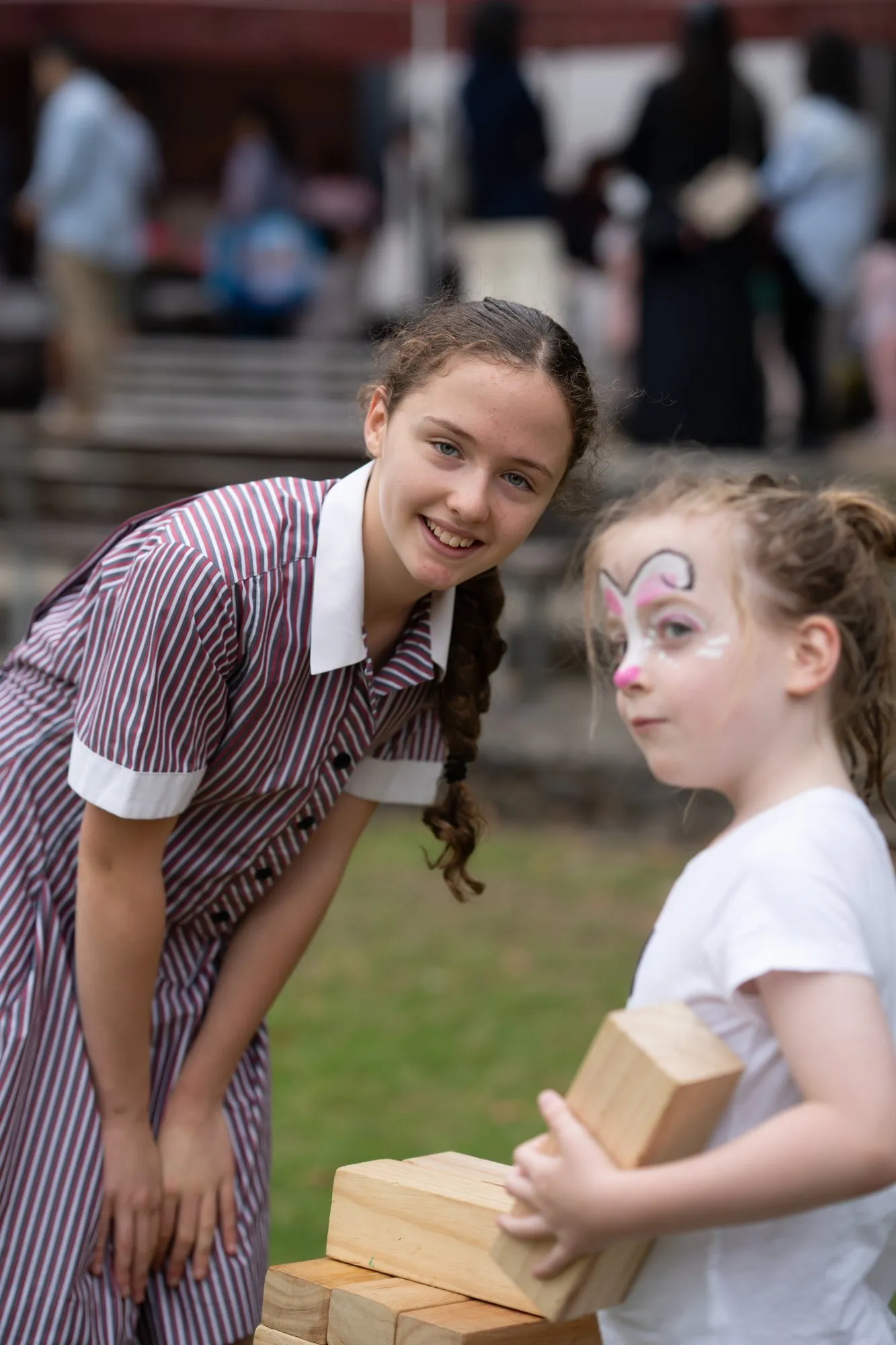 Smiling girl in a striped dress leans forward near a young child with face paint holding wooden blocks outdoors.