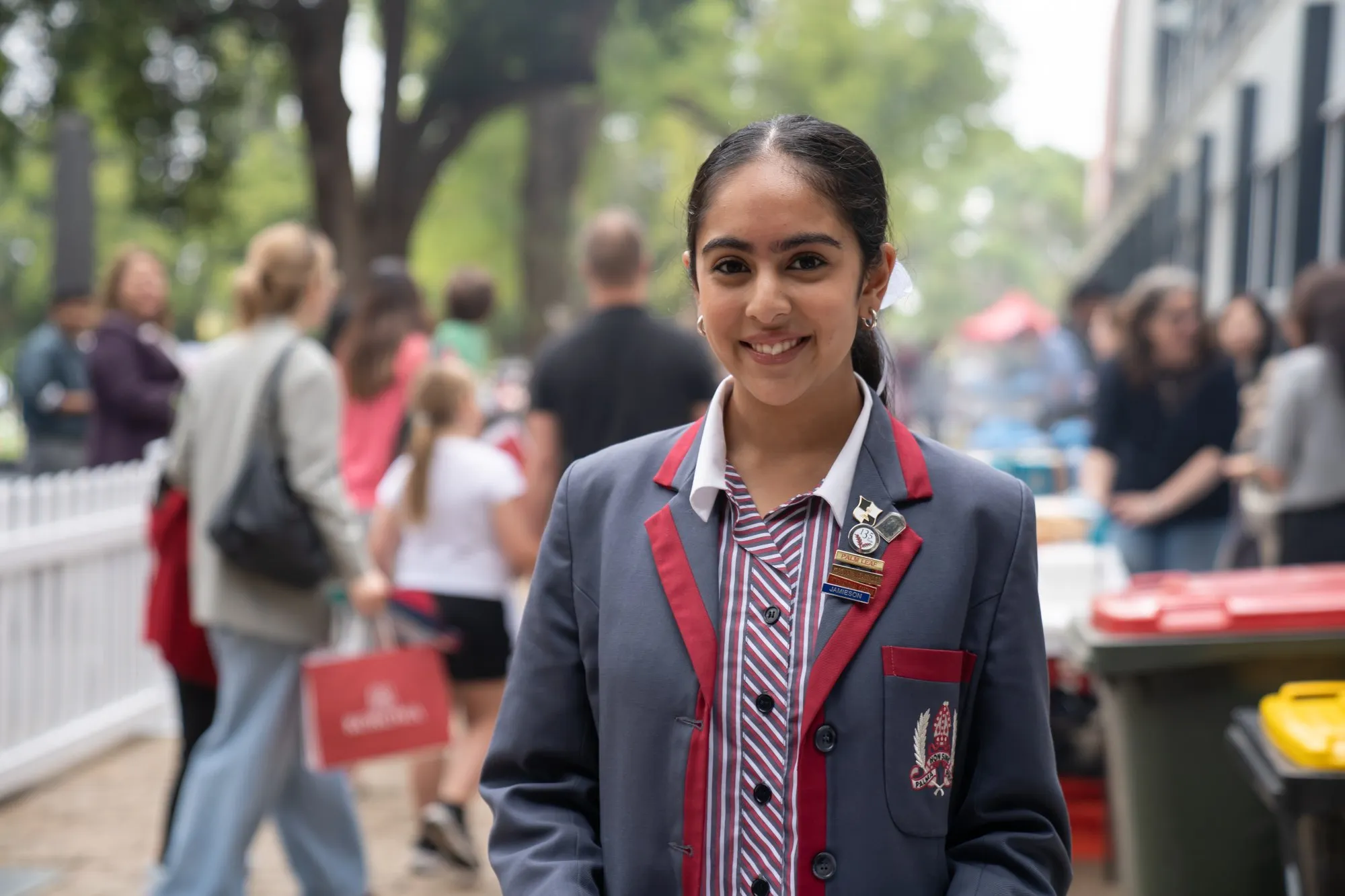 Smiling girl in a school uniform with red and blue accents standing outdoors with blurred people in the background.