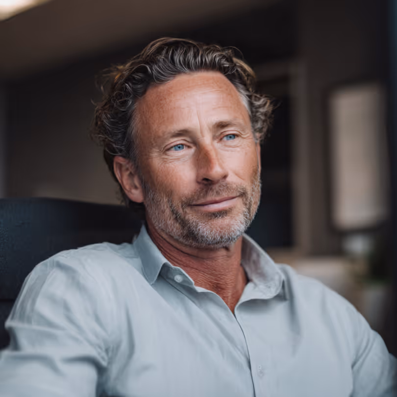 Middle-aged man with curly hair and beard wearing a light blue shirt, sitting and looking thoughtfully.