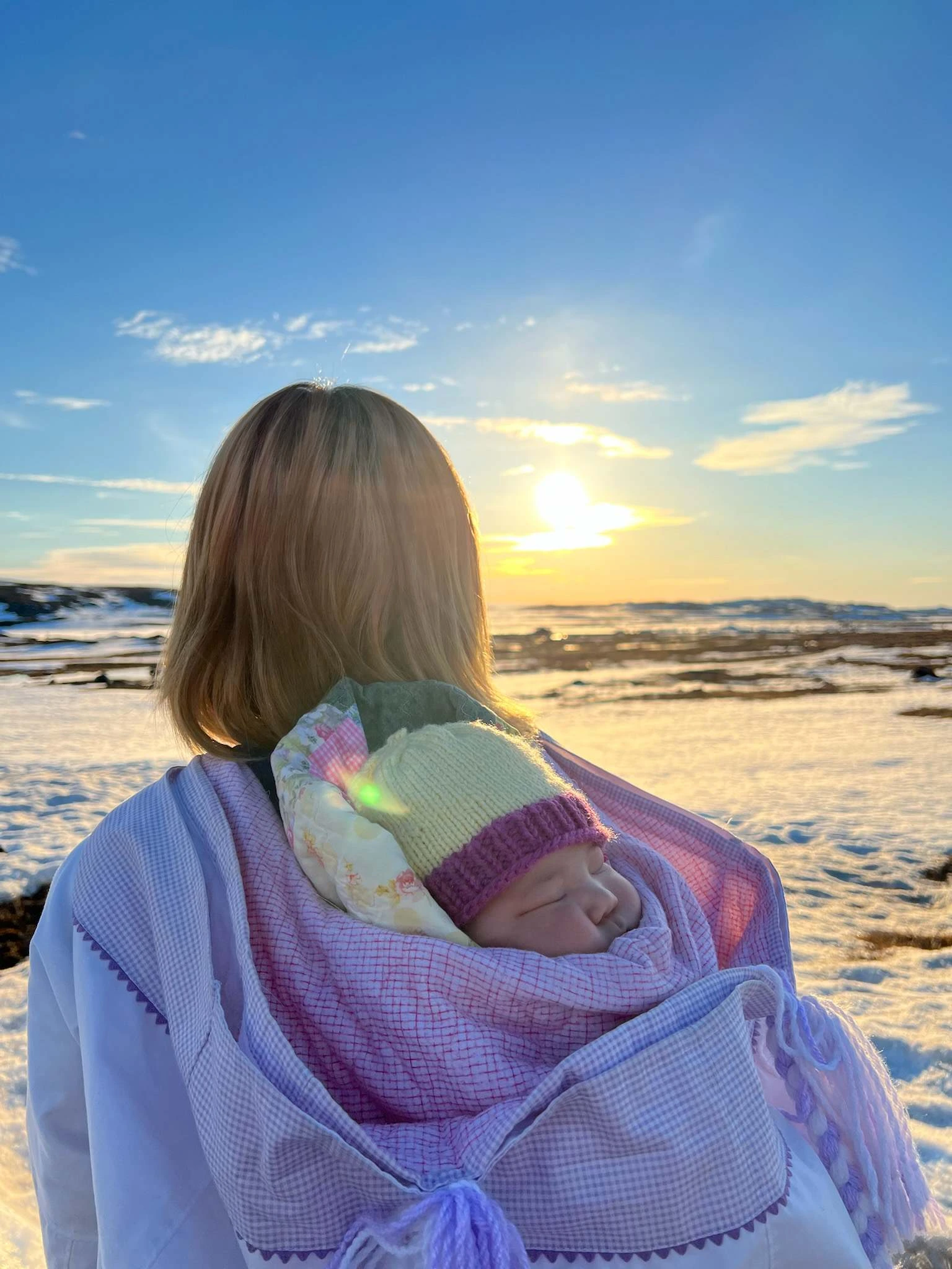 A woman with a sleeping infant on her back looks out over the sunset.