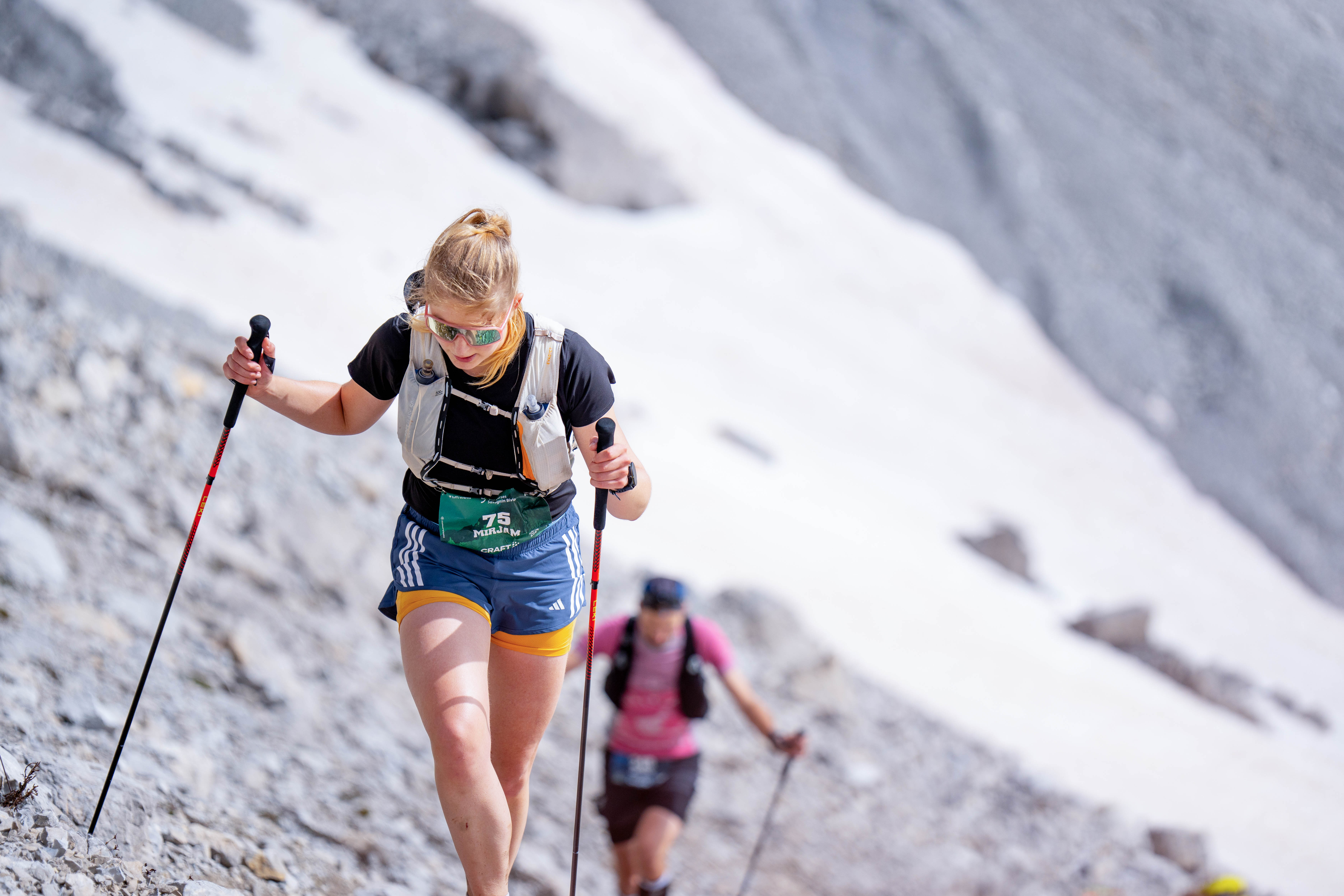 Zwei Bergläufer mit Trekkingstöcken steigen auf steinigem Pfad mit Schneefeldern im Hintergrund auf.