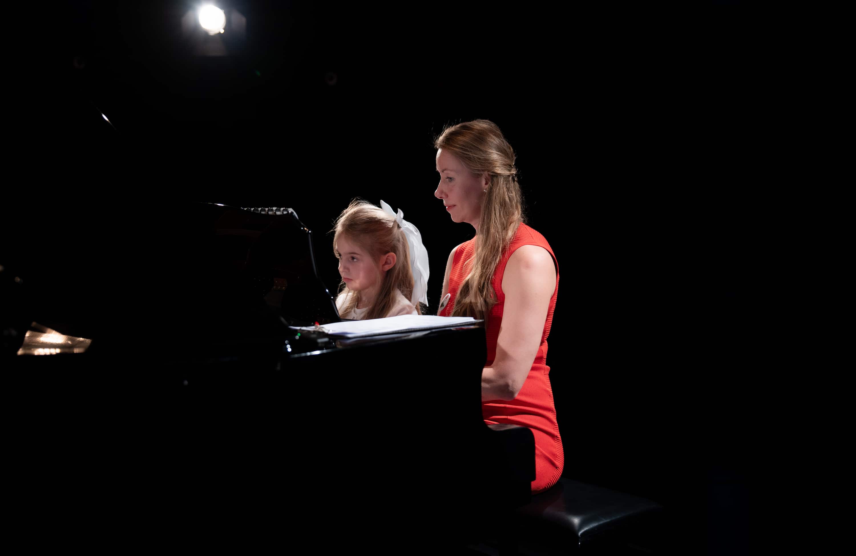 Zwei Frauen sitzen nebeneinander am schwarzen Flügelklavier, eine trägt ein rotes ärmelloses Kleid, die andere ein weisses Oberteil.