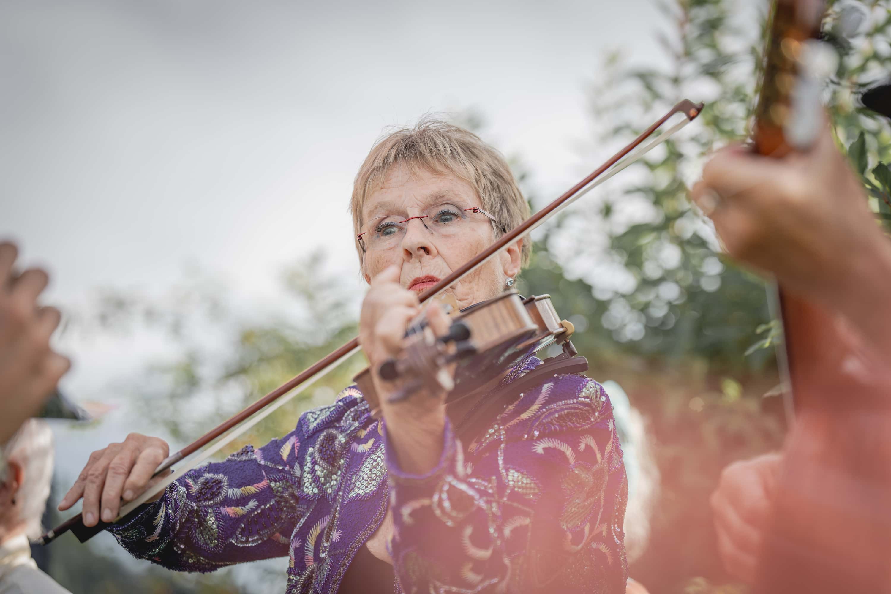 Person in blauem, gemustertem Oberteil spielt Geige, umgeben von weiteren Musikern mit Streichinstrumenten.