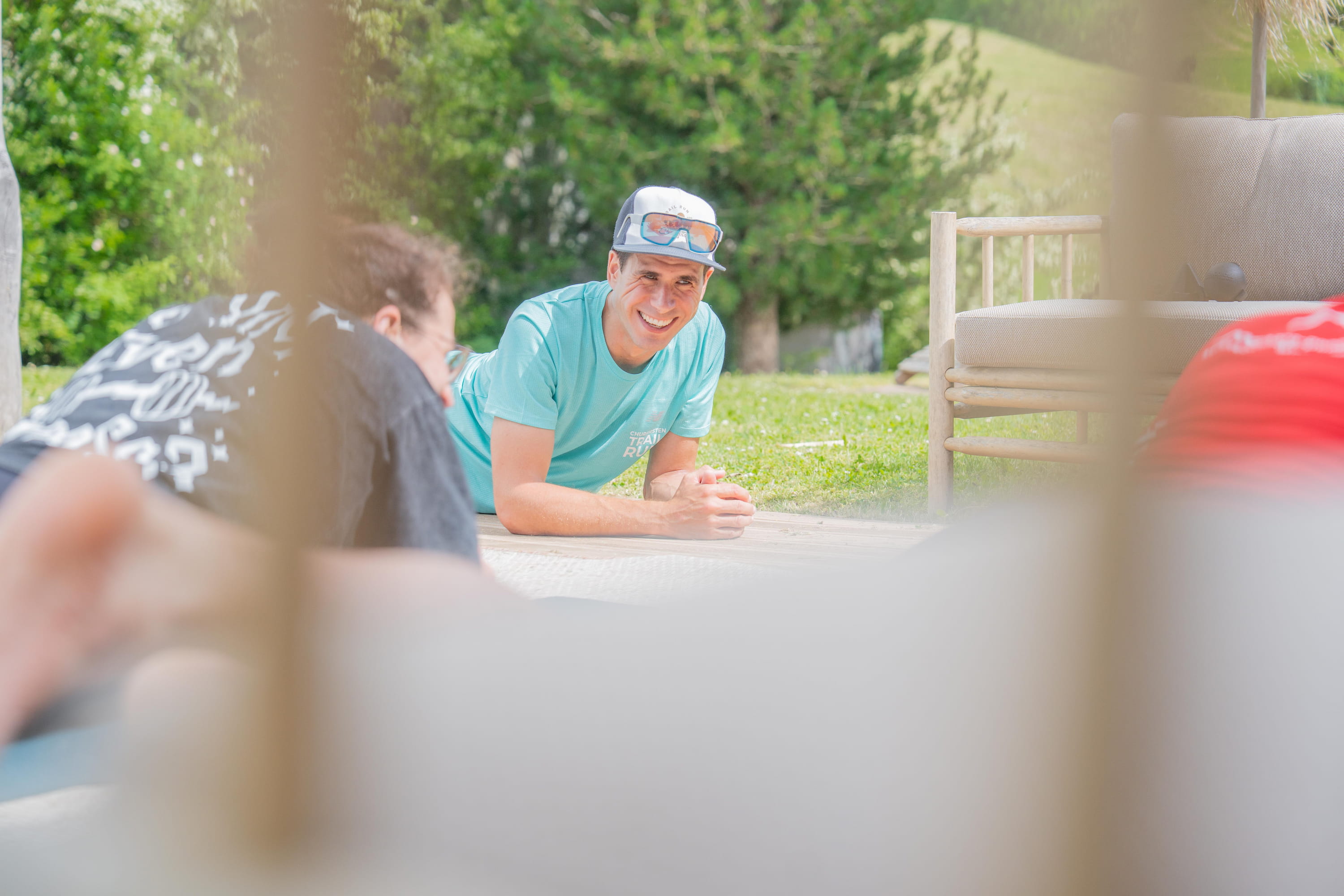 Zwei Männer liegen auf dem Boden im Freien, einer trägt ein hellblaues T-Shirt und eine weiße Kappe, im Hintergrund grüne Bäume und eine Gartenbank. Sie machen Blackroll Übungen.