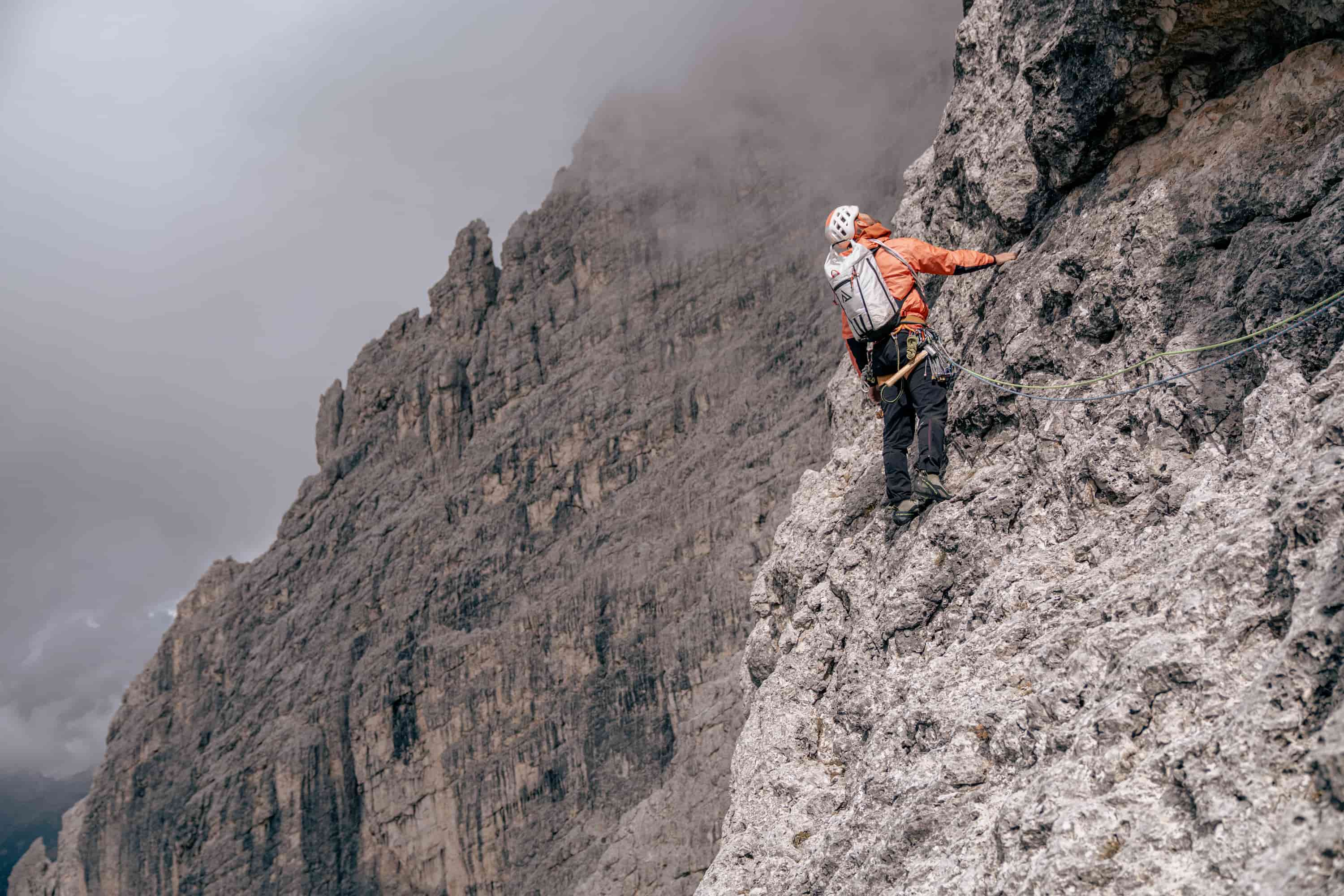 Kletterer in orange Jacke und weißem Helm an steiler Felswand mit grauen, wolkenverhangenen Bergen im Hintergrund.