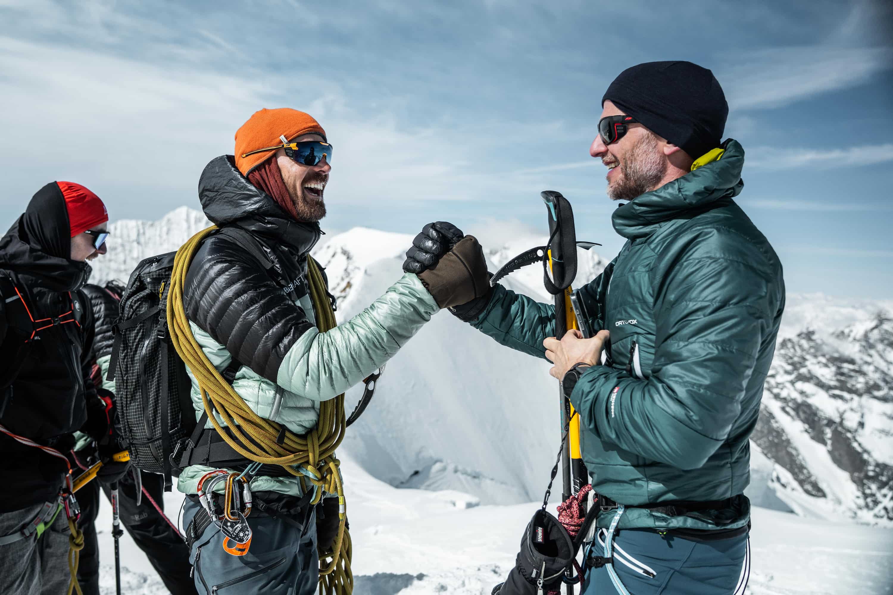 Zwei Bergsteiger in Winterkleidung und mit Kletterausrüstung geben sich auf schneebedecktem Berggipfel die Faust.