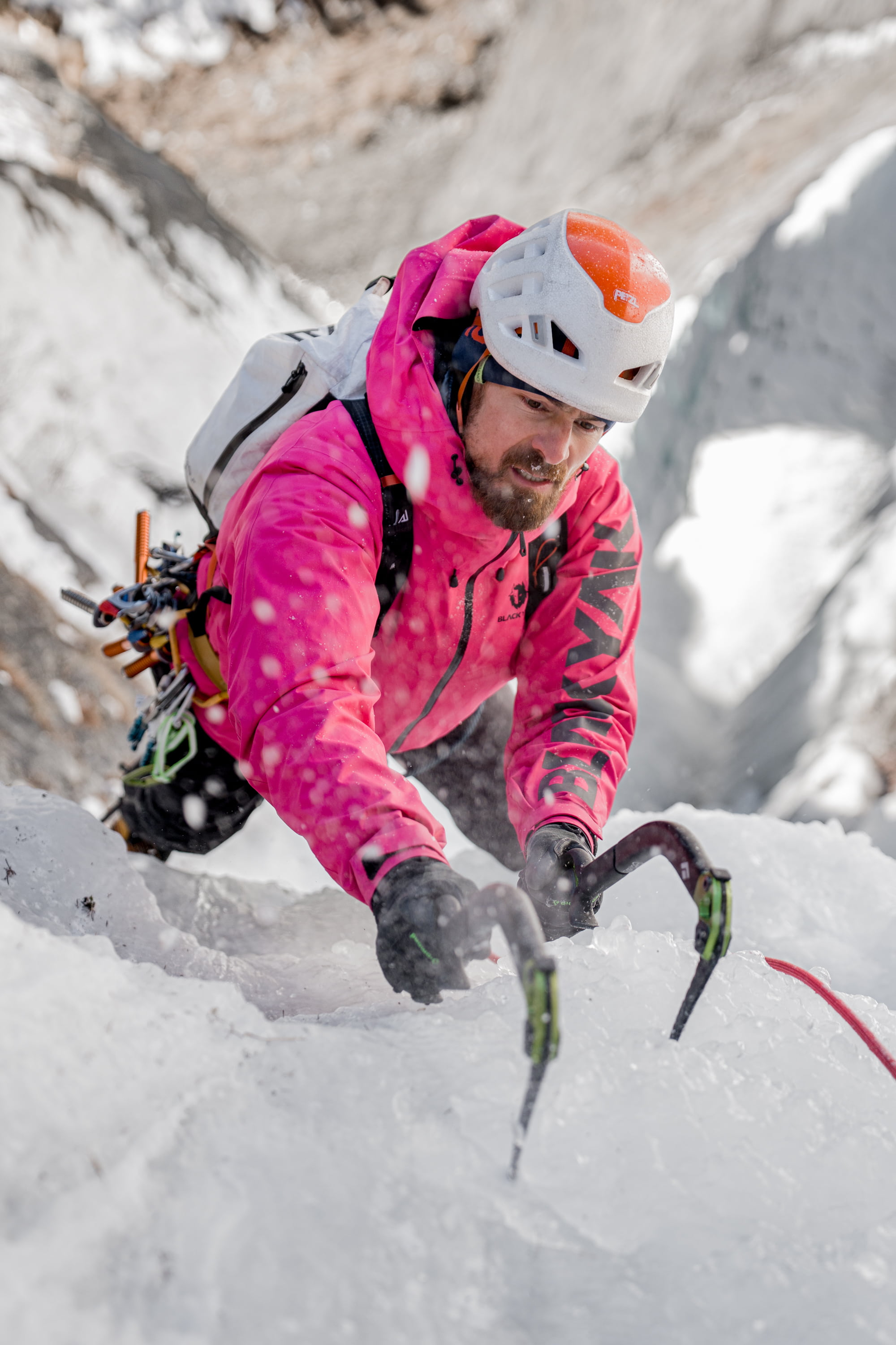 Person in pinkfarbener Jacke und weißem Helm klettert mit Eispickeln an einer steilen Eiswand.