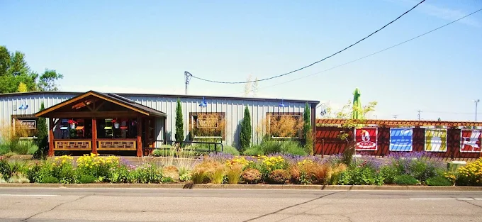 Outdoor garden center with a wooden gazebo, various plants and flowers, and a metal building in the background under a clear blue sky.
