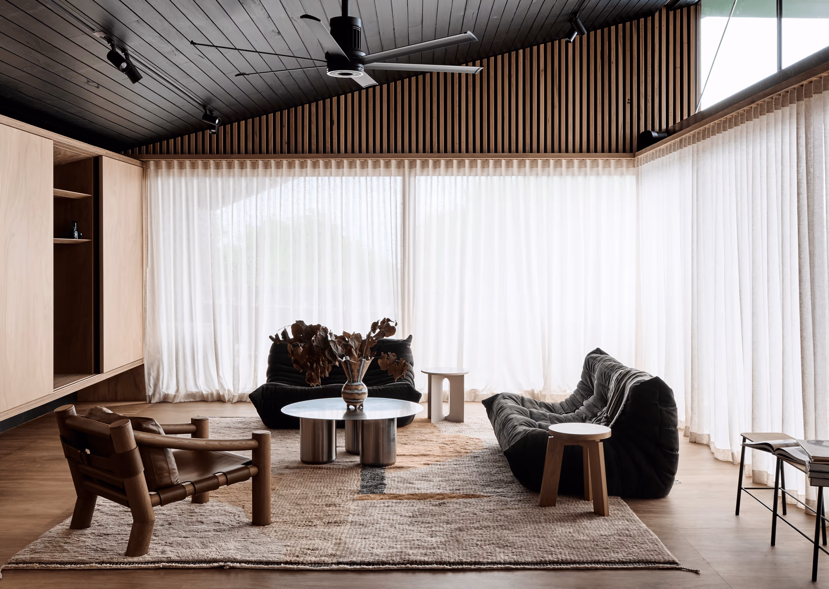 Modern minimalist living room with wooden furniture, black sofas, and large sheer curtains.