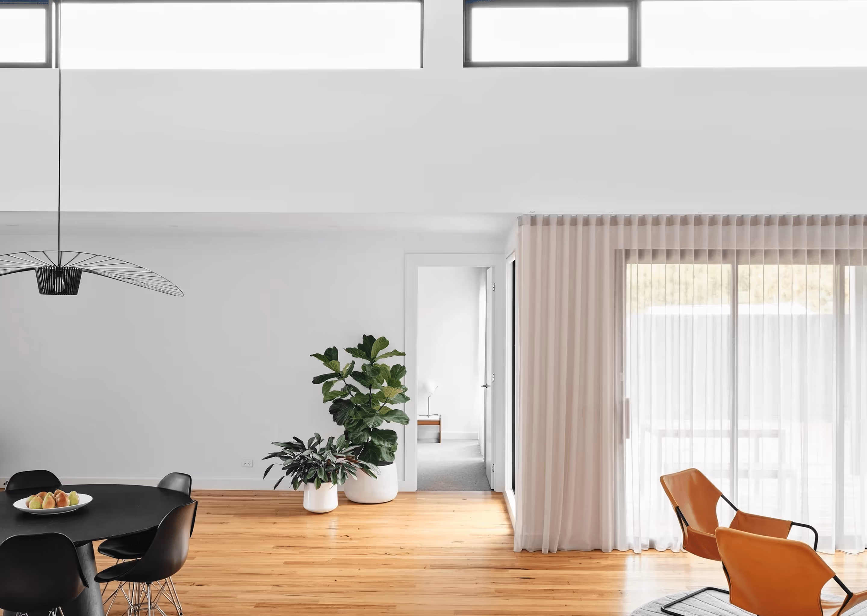 Bright, modern dining and living area with wooden floors, black dining chairs, leather lounge chairs, and large windows with sheer curtains.