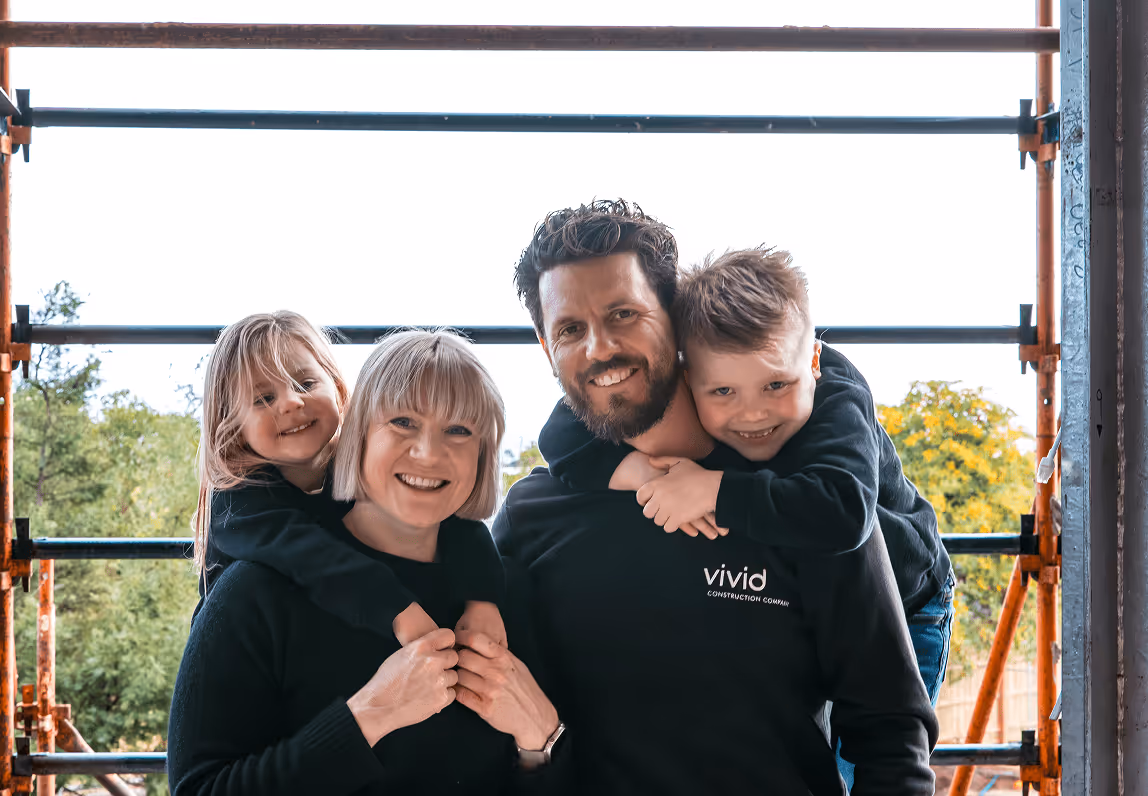 Family of four smiling, outdoors, with children on parents' backs.