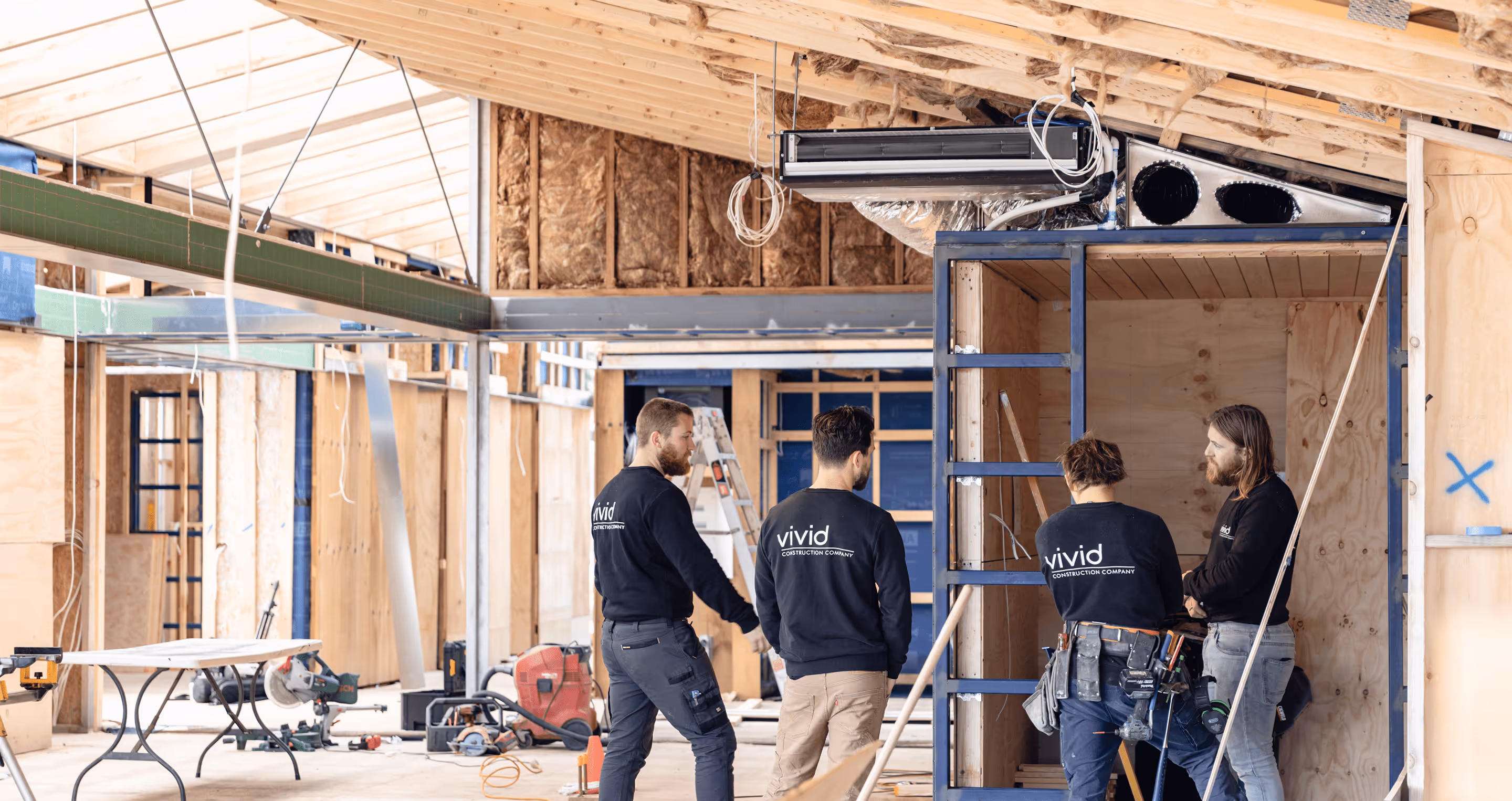 Four construction workers discussing plans inside a partially built wooden structure.