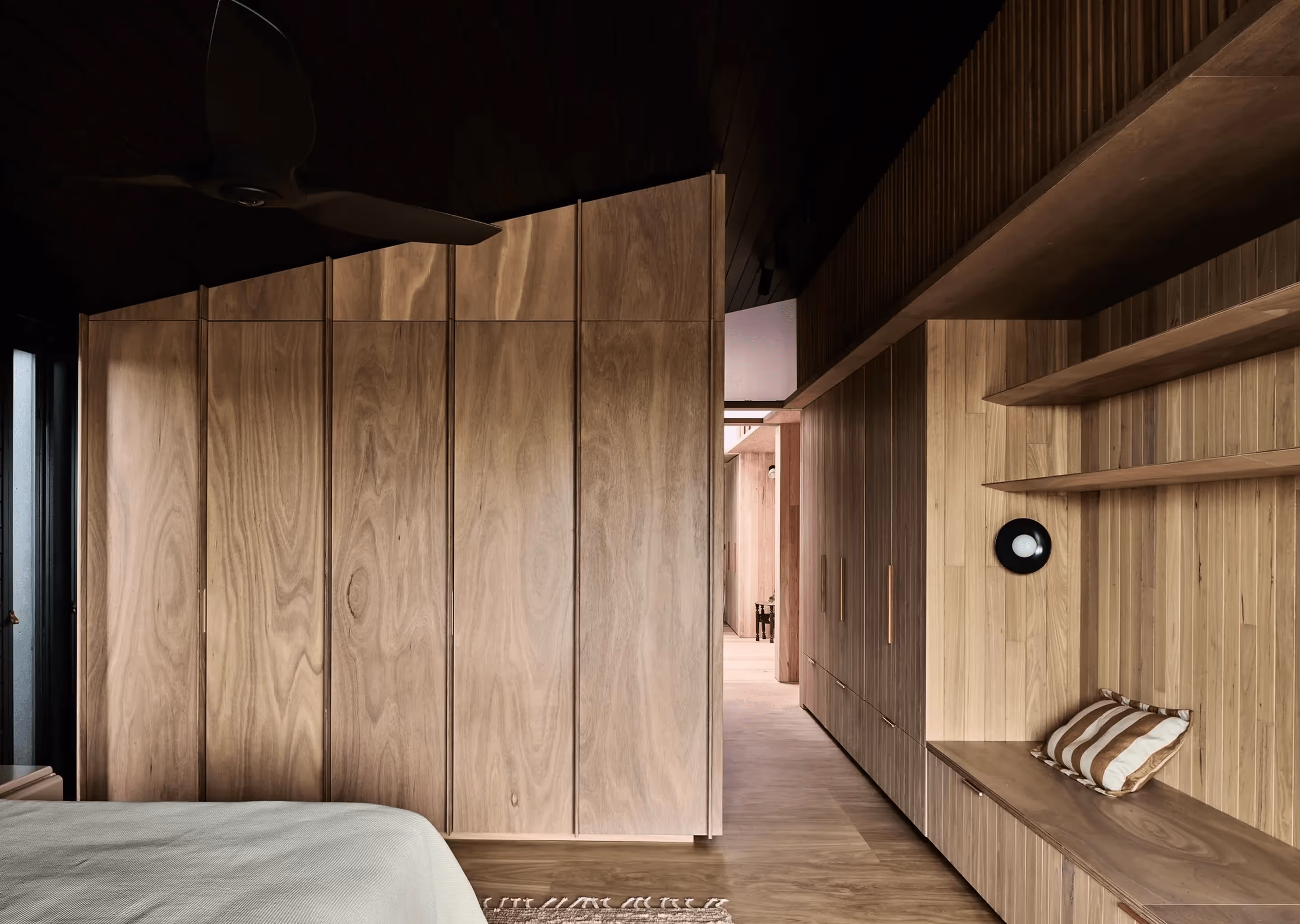Minimalist wooden bedroom interior with built-in wardrobes, shelves, and a bench with a striped cushion.