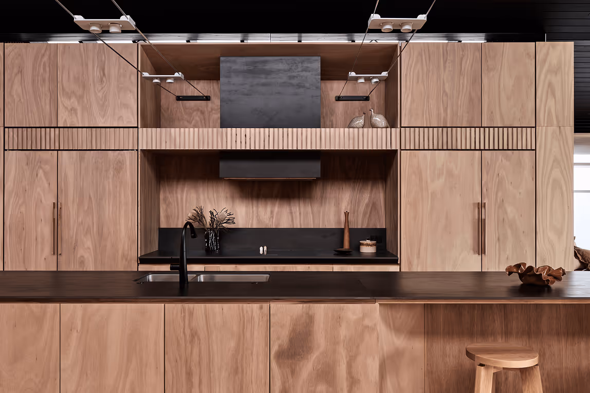 Modern kitchen with natural wood cabinetry, black countertop, built-in stove area, and a central island with a sink and wooden stool.