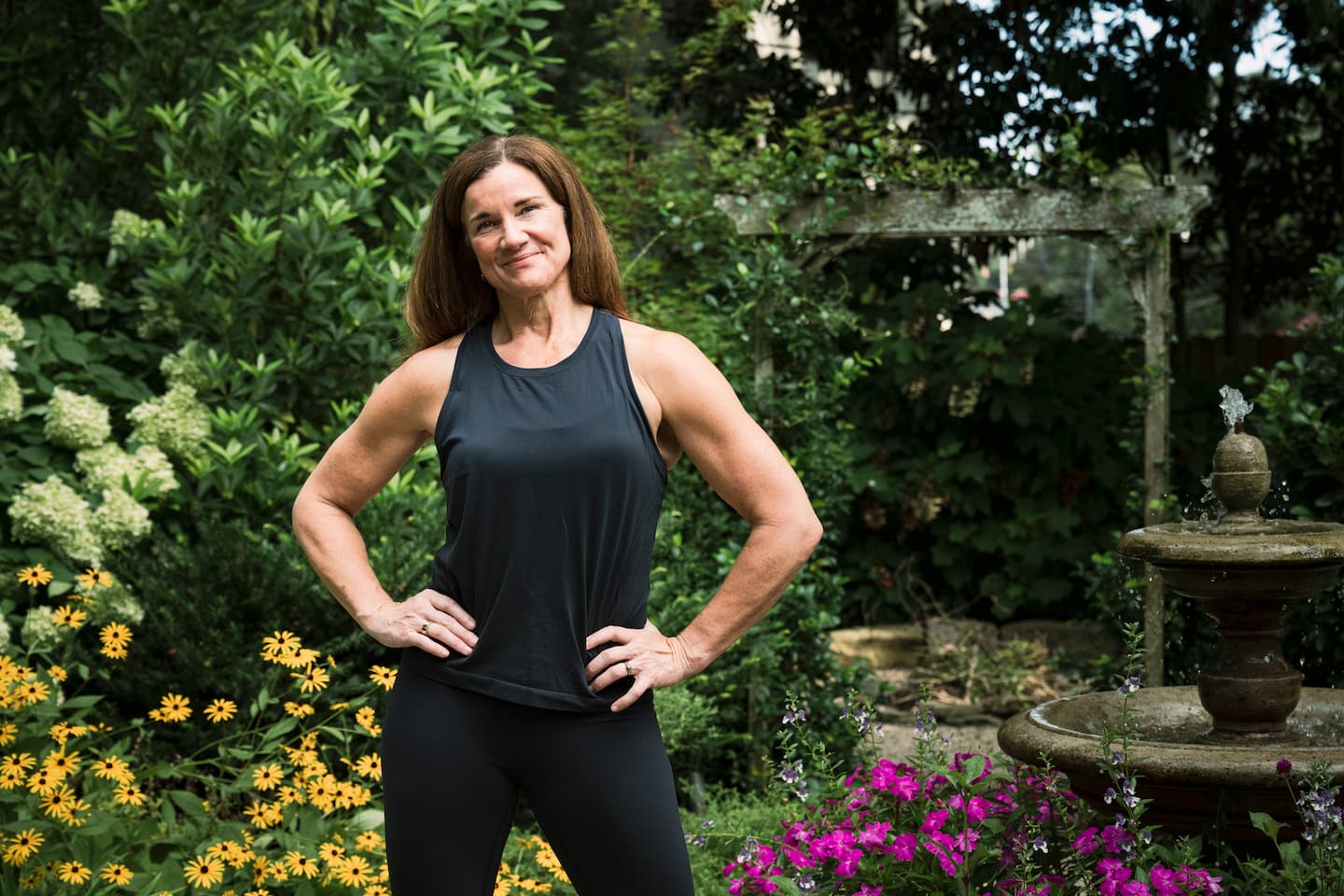 A fit middle-aged woman stands confidently outside in a garden.
