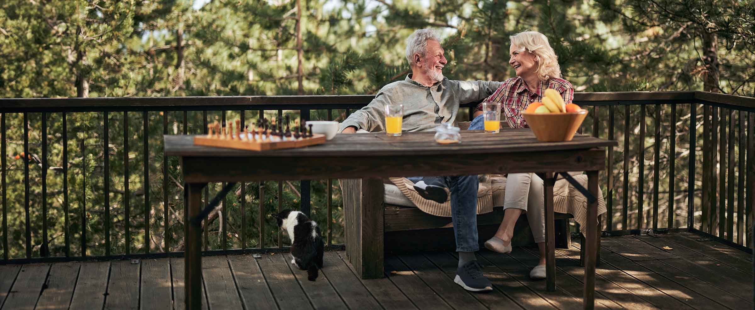 Smiling senior couple sitting on a wooden deck with drinks, a fruit bowl, and a chessboard, enjoying nature with a cat nearby
