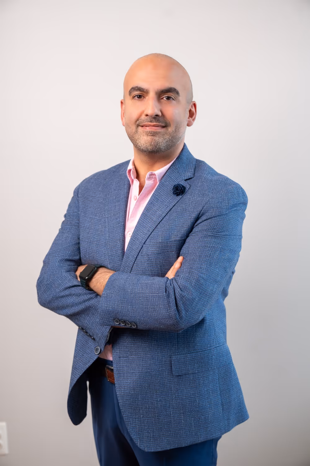 Confident bald man wearing a blue blazer and pink shirt with arms crossed, standing against a plain light background.