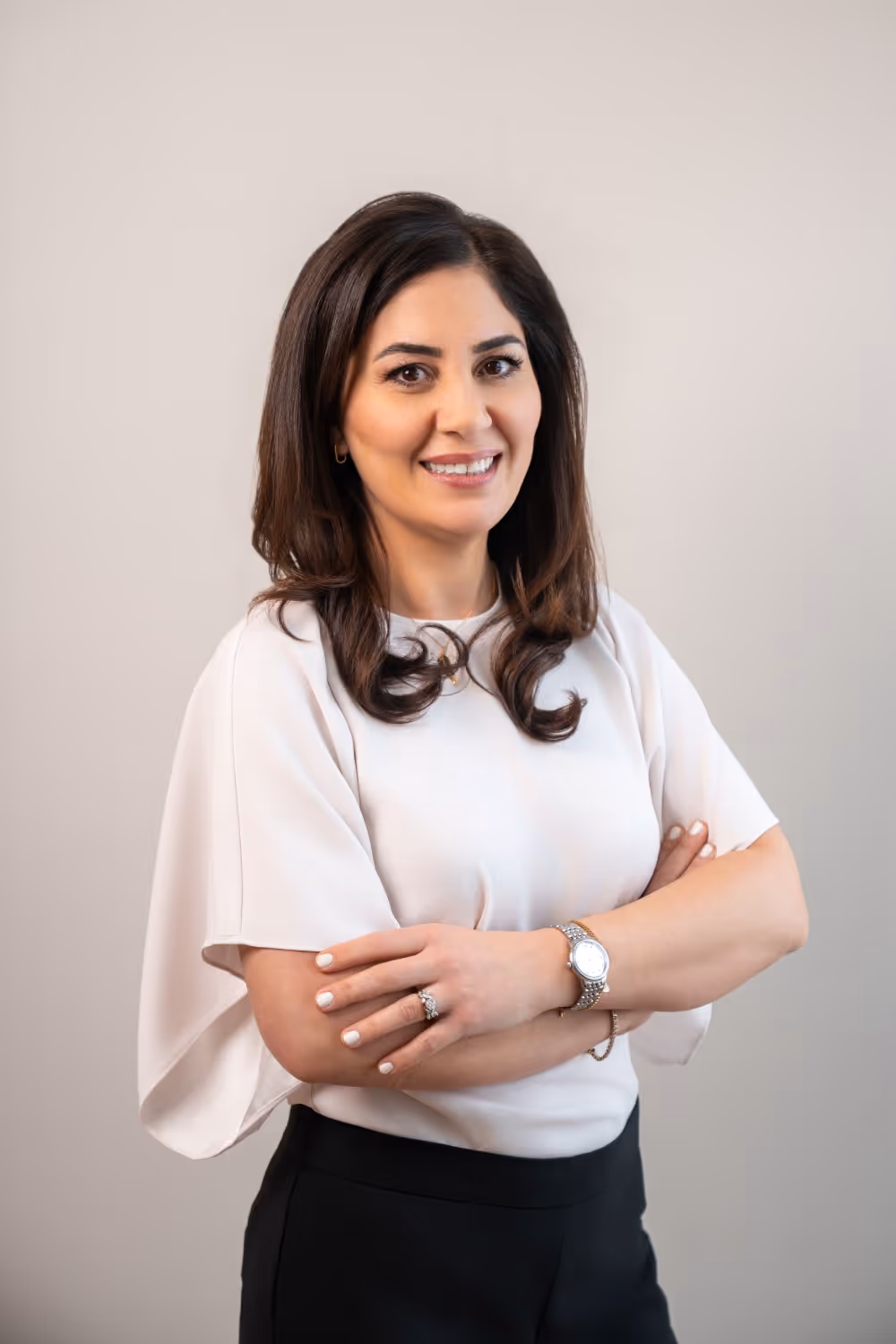 Smiling woman with dark hair wearing a white blouse and black pants, arms crossed, against a plain background.