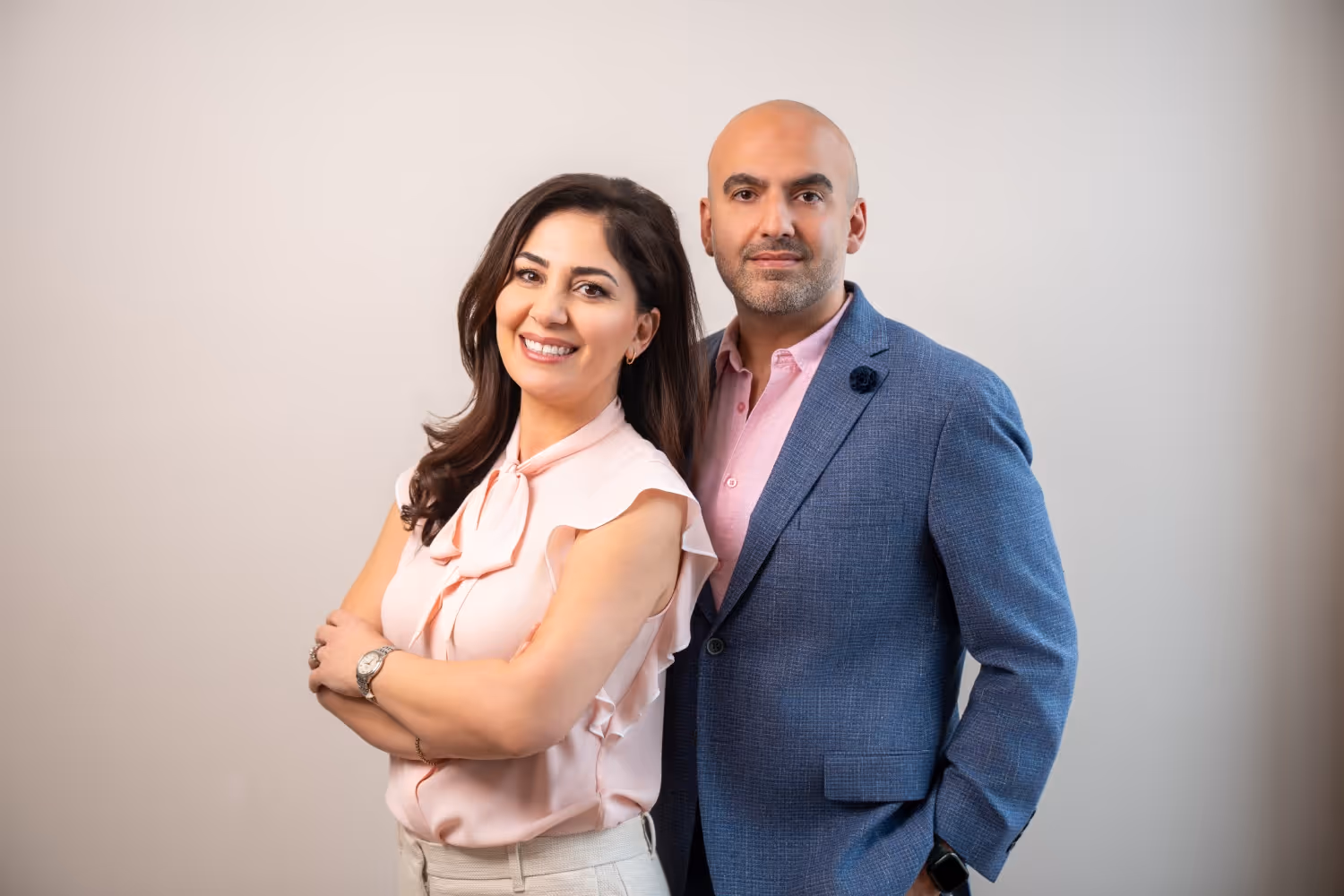 Smiling man in blue blazer and woman in light pink blouse standing side by side against a plain background.
