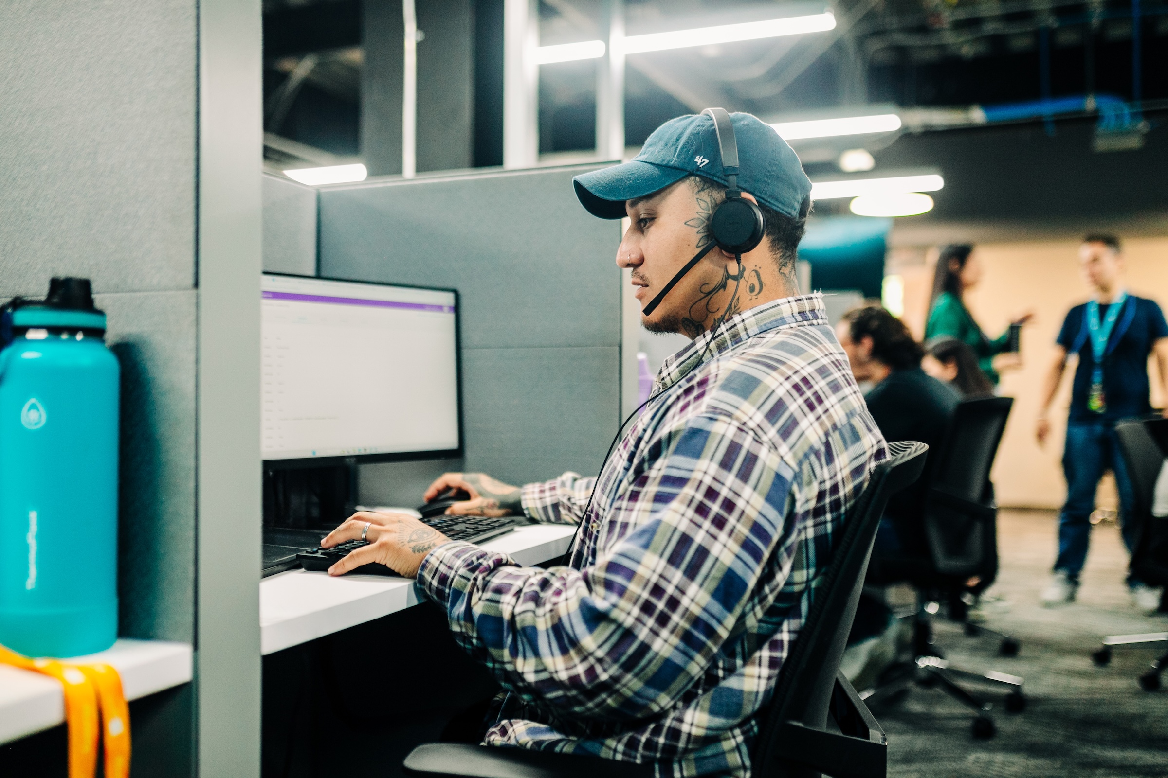 Man with tattoos wearing a headset and plaid shirt working on a computer in an open office space.