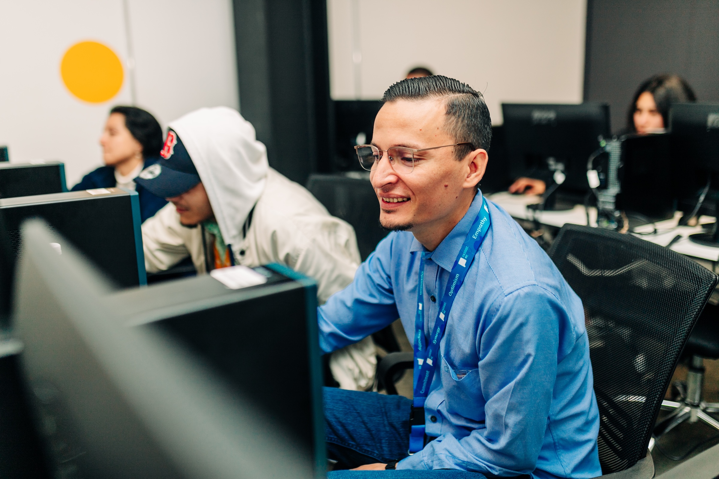 Man wearing glasses and a blue shirt working on a computer in an office with colleagues.