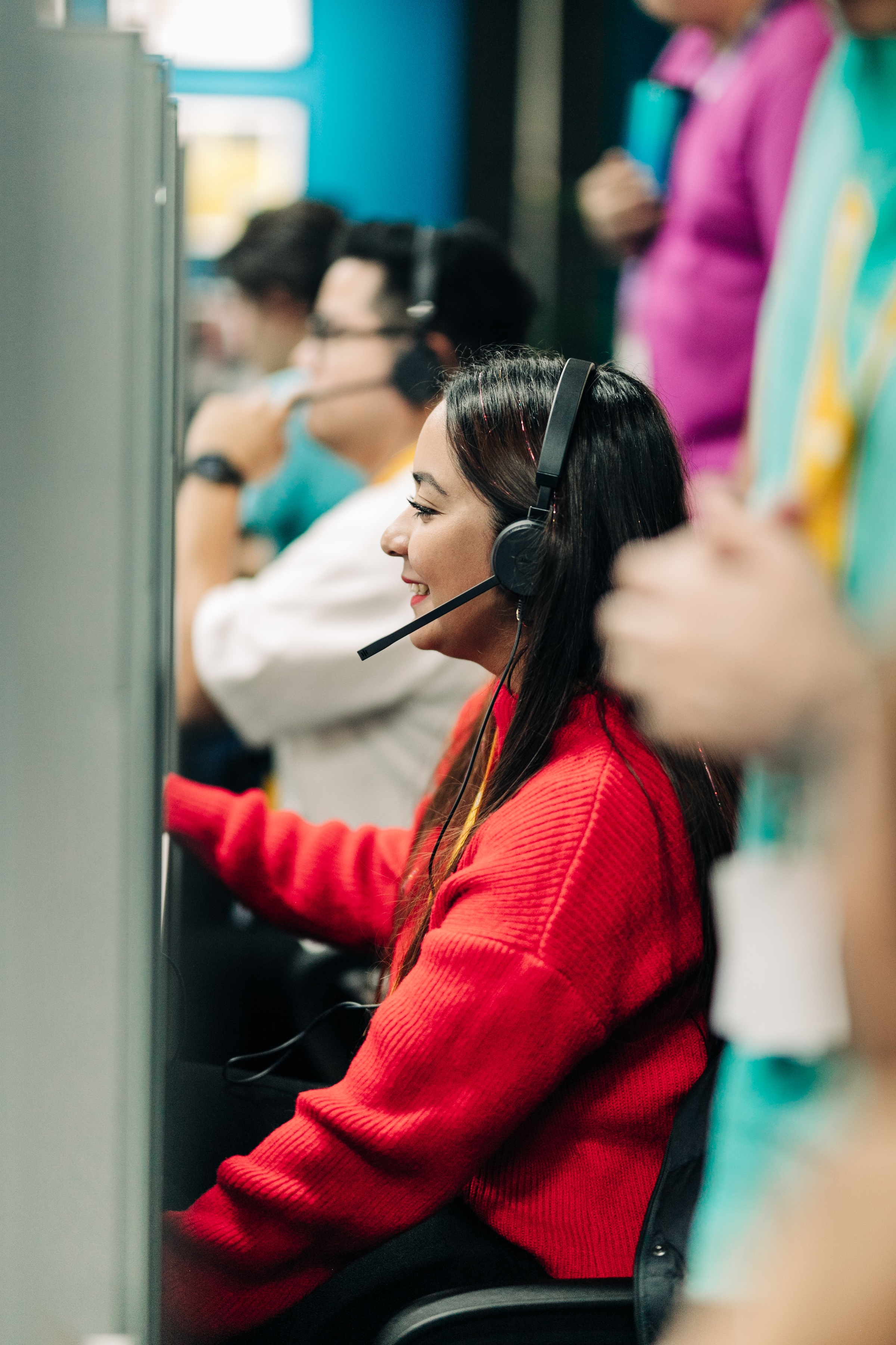 Woman wearing a red sweater and headset working at a computer station in a busy call center.